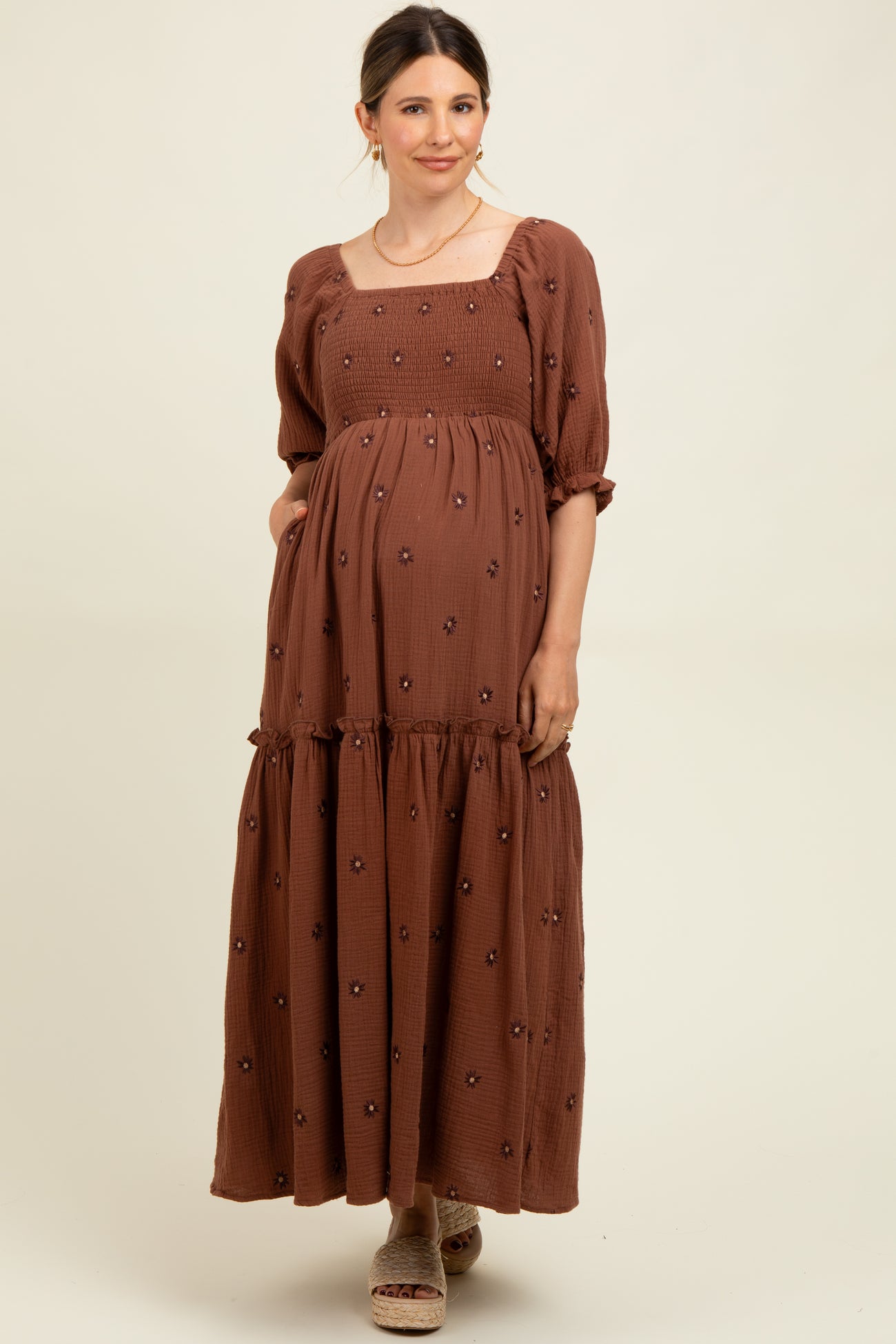 woman wearing a brown smocked maternity maxi dress with puff sleeves and floral embroidery, holding her baby bump