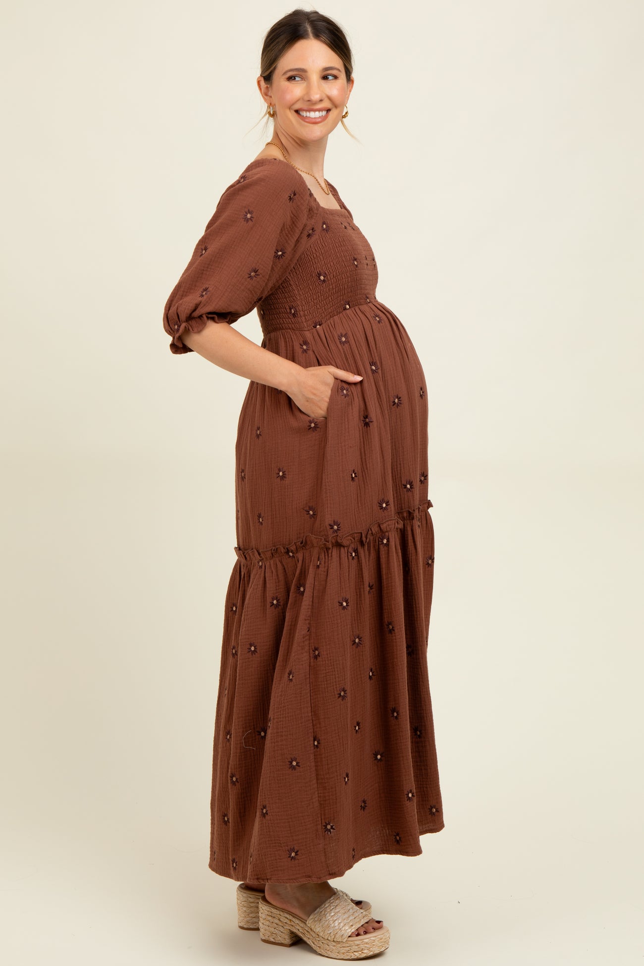 woman wearing a brown maternity maxi dress with puff sleeves and floral embroidery, showing her baby bump and pockets