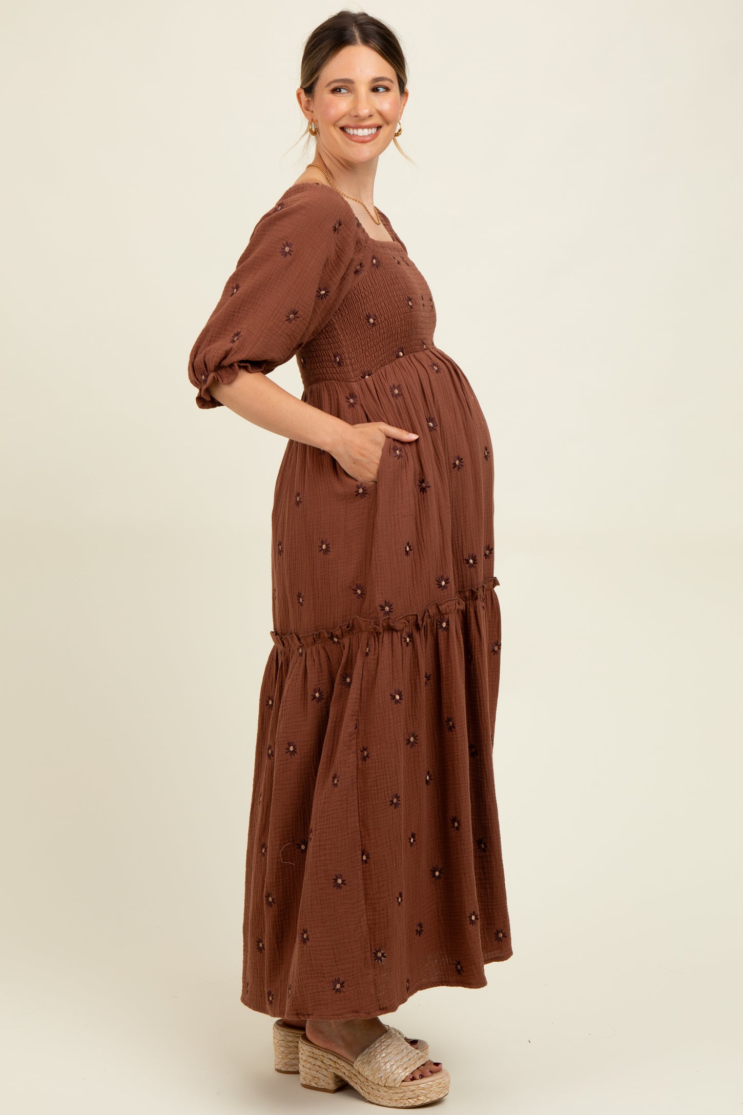 woman wearing a brown maternity maxi dress with puff sleeves and floral embroidery, showing her baby bump and pockets