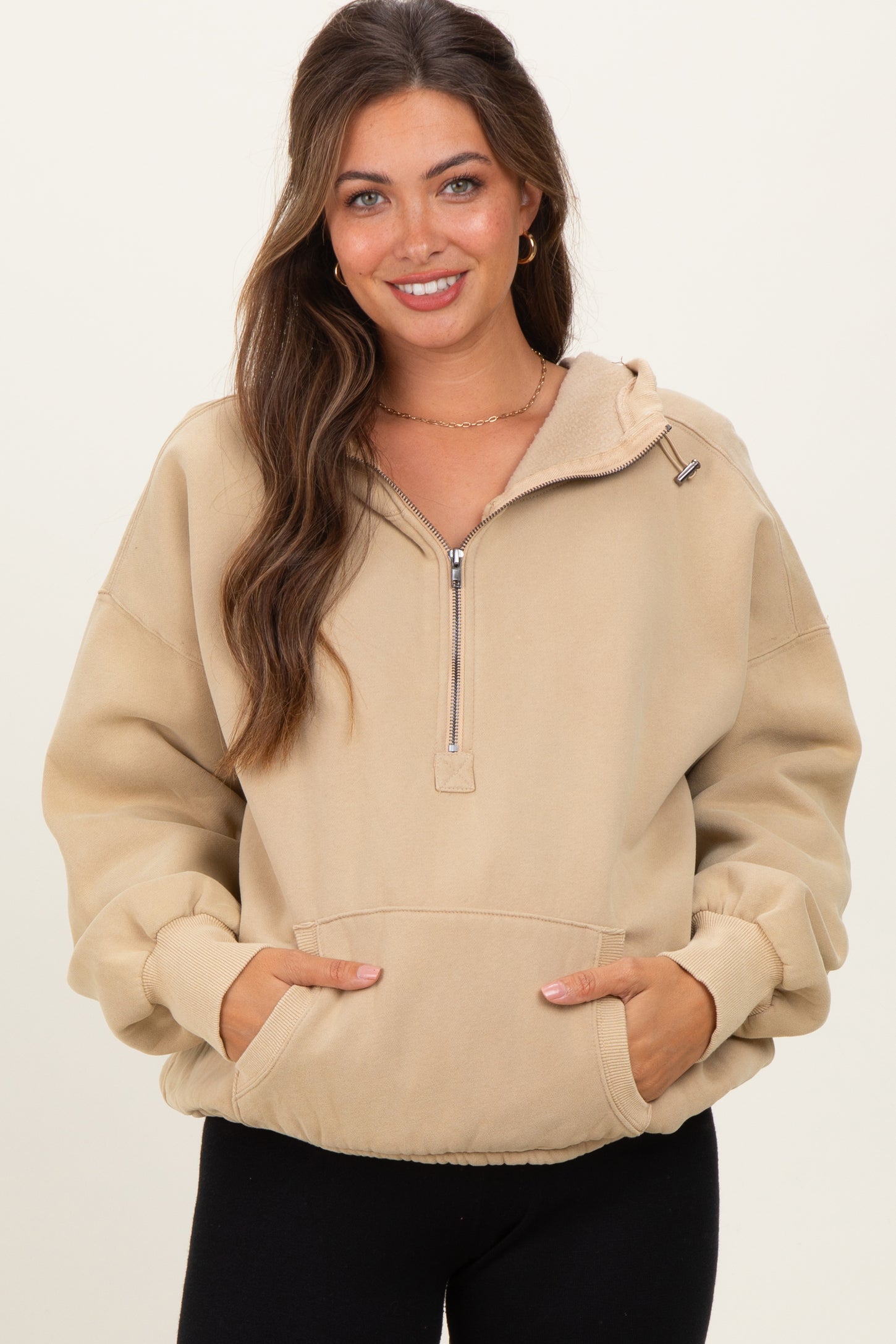 woman wearing a beige maternity hoodie with a zippered neck and front pockets, smiling at the camera