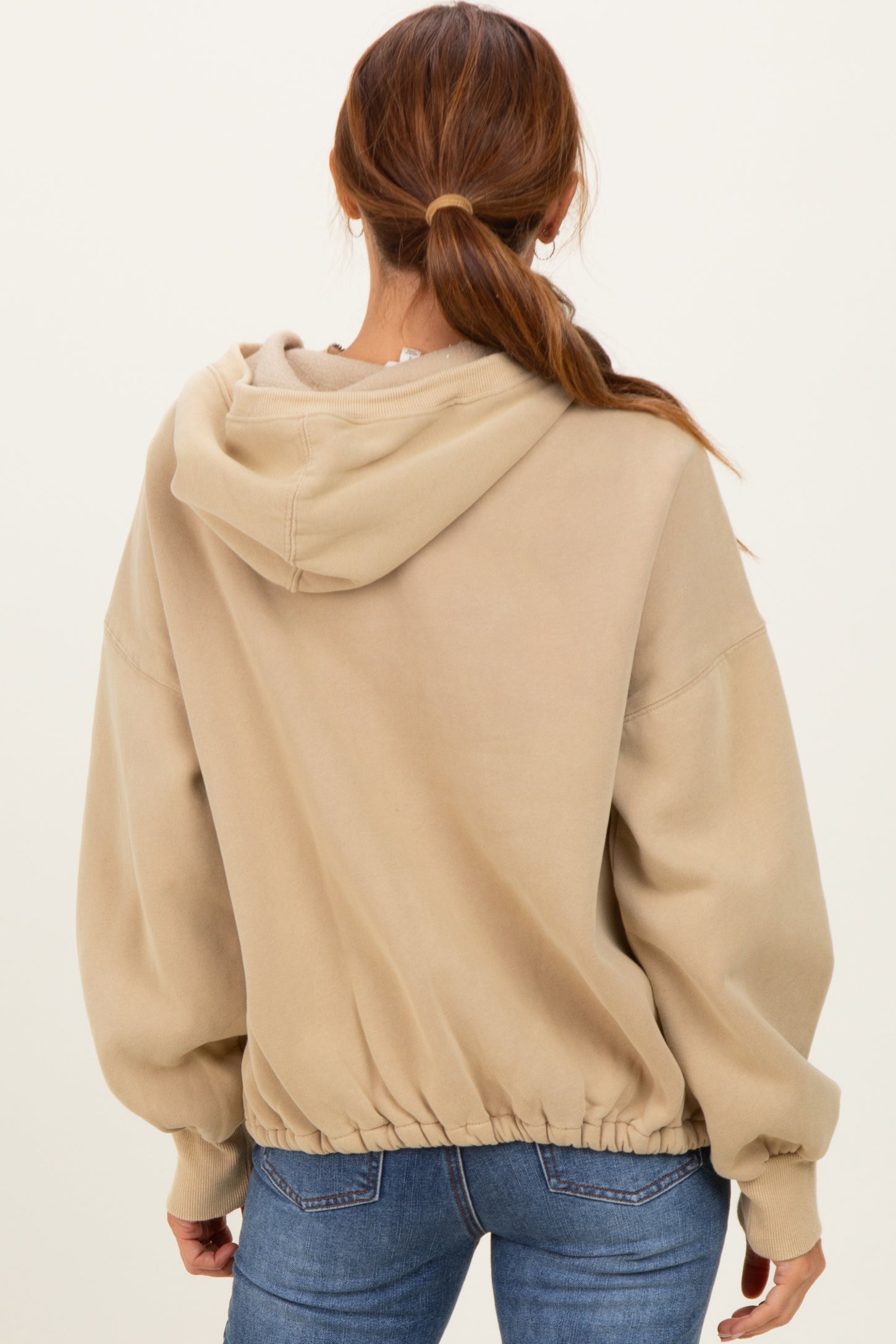 woman wearing a beige oversized hoodie with a cinched hem and a hood, viewed from the back