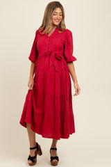 woman wearing a red maternity dress with short puff sleeves, tie waist, and tiered design, showing her baby bump