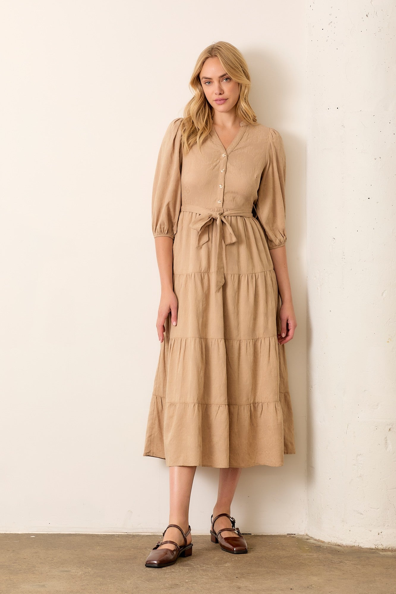 woman wearing a tan tiered midi dress with puff sleeves and a tie waist, standing against a wall