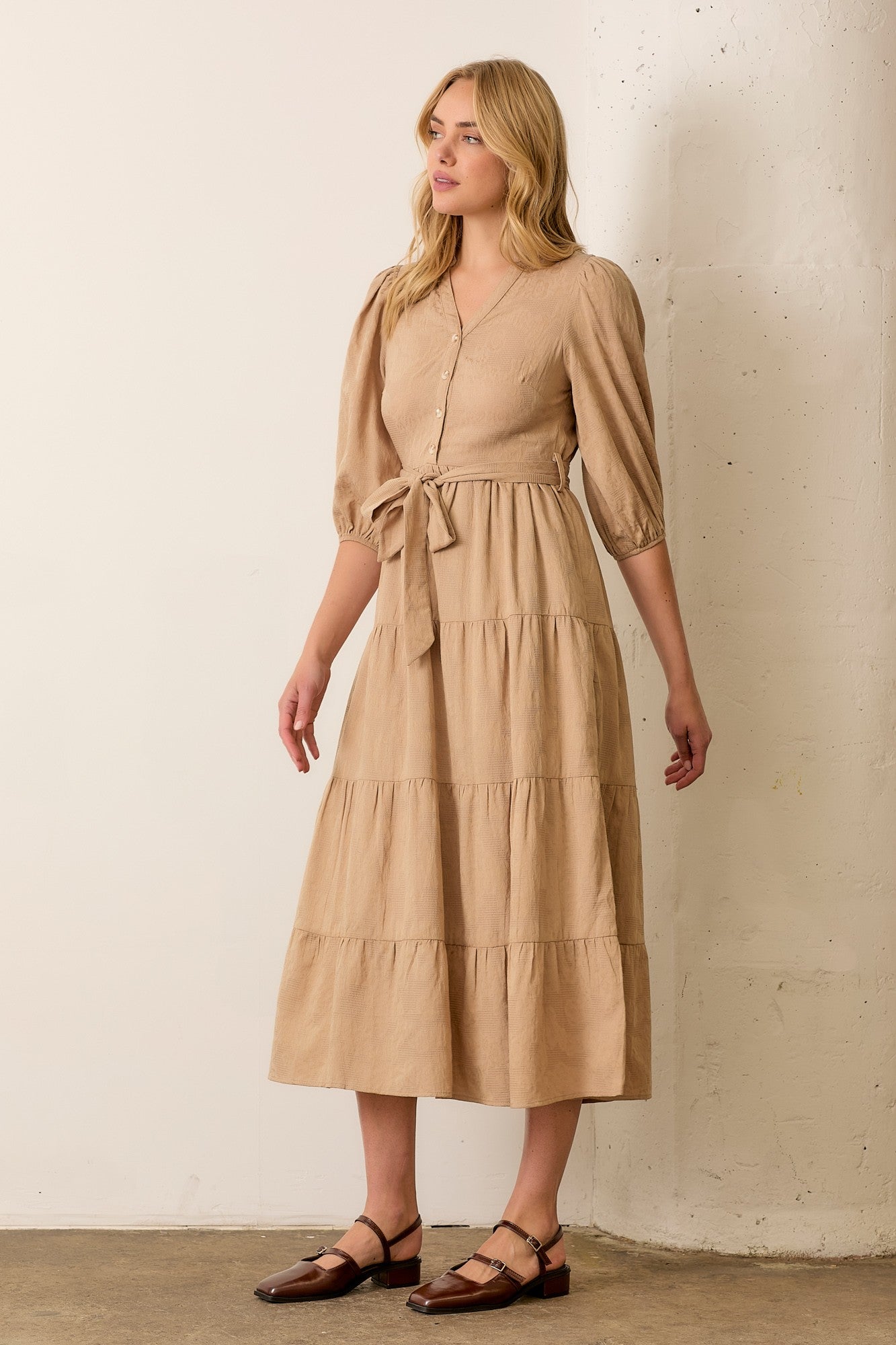 woman wearing a beige midi dress with puff sleeves and a tie waist detail, standing against a neutral backdrop