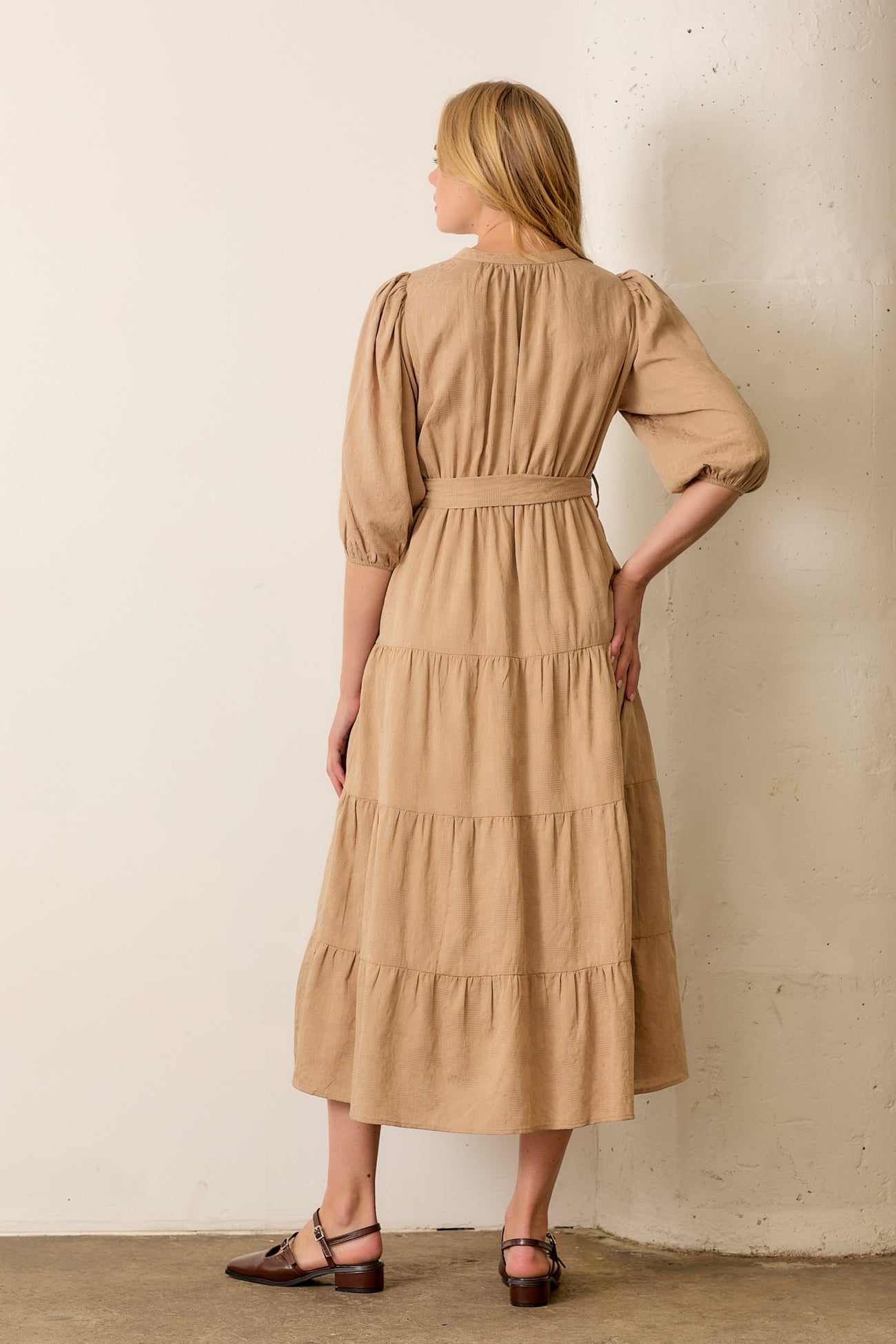 woman wearing a tan tiered midi dress with puff sleeves and a belted waist, standing with her back to the camera