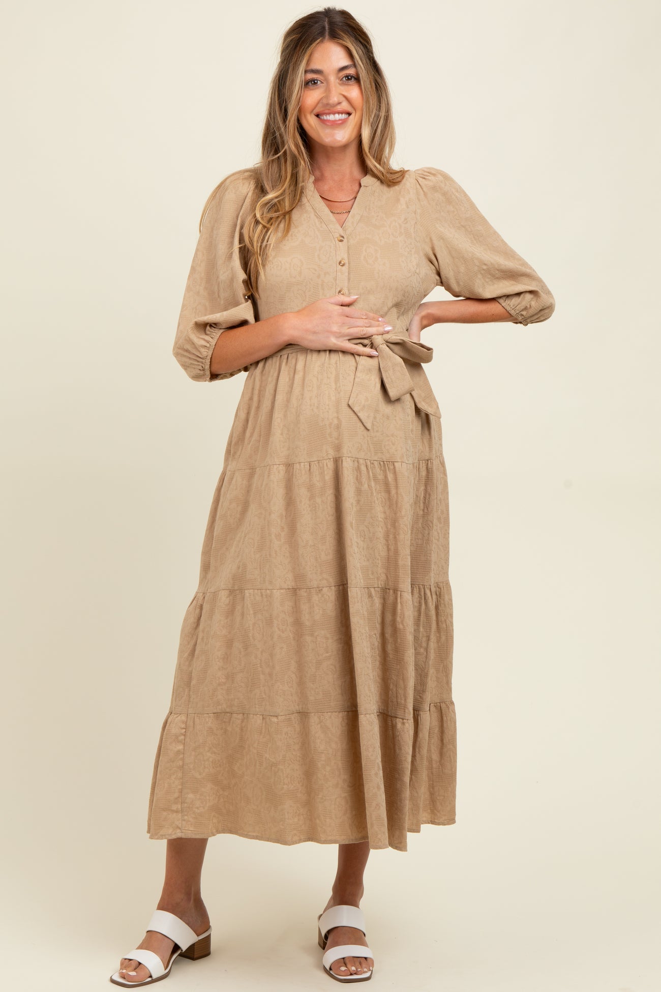 woman wearing a tan maternity midi dress with button details and a tie waist, showcasing her baby bump