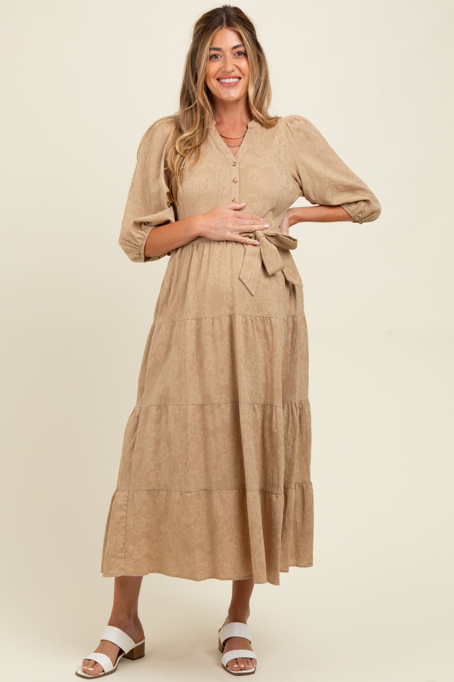 woman wearing a tan maternity midi dress with button details and a tie waist, showcasing her baby bump