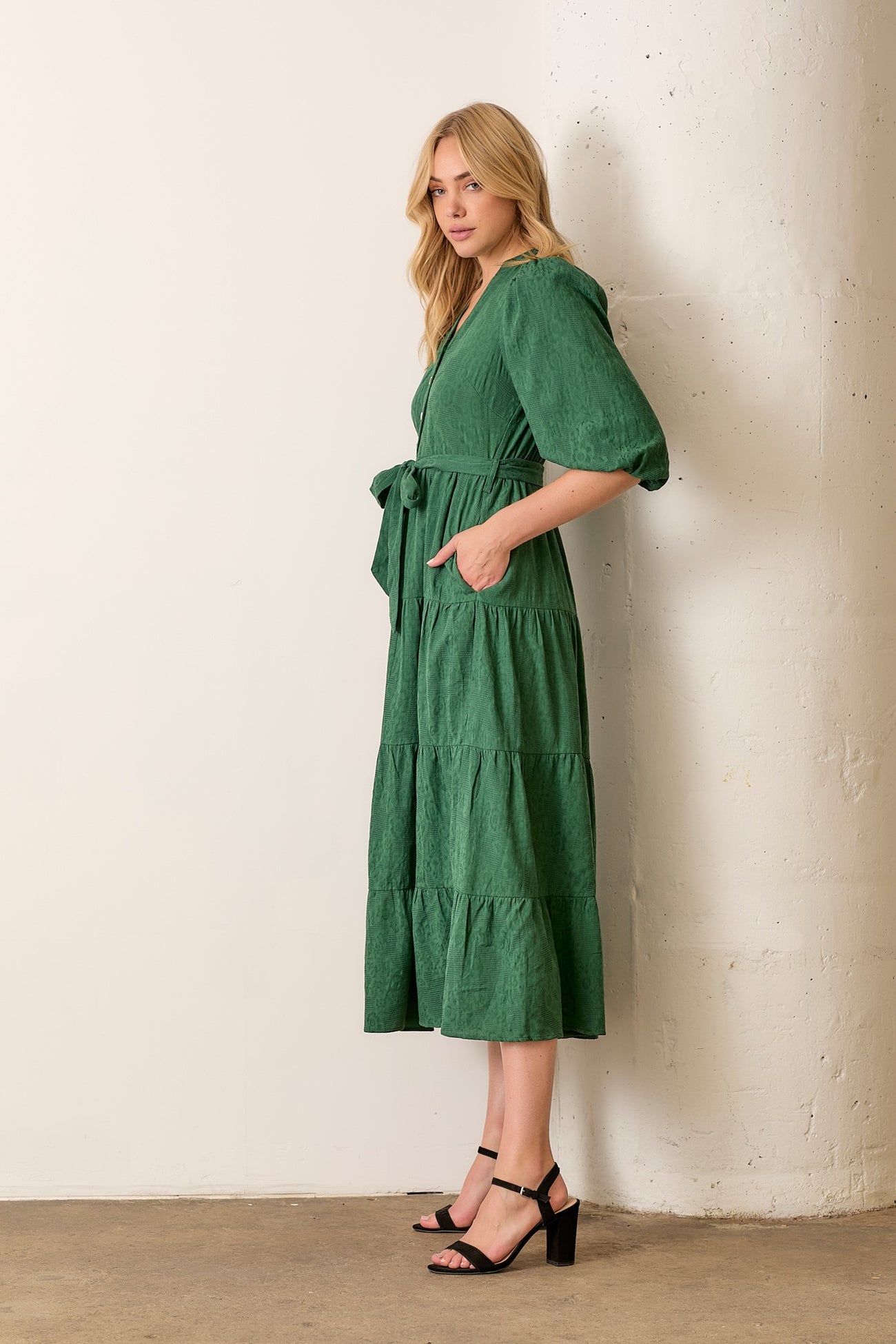 woman wearing a green tiered midi dress with puff sleeves and a tie waist, standing against a white wall