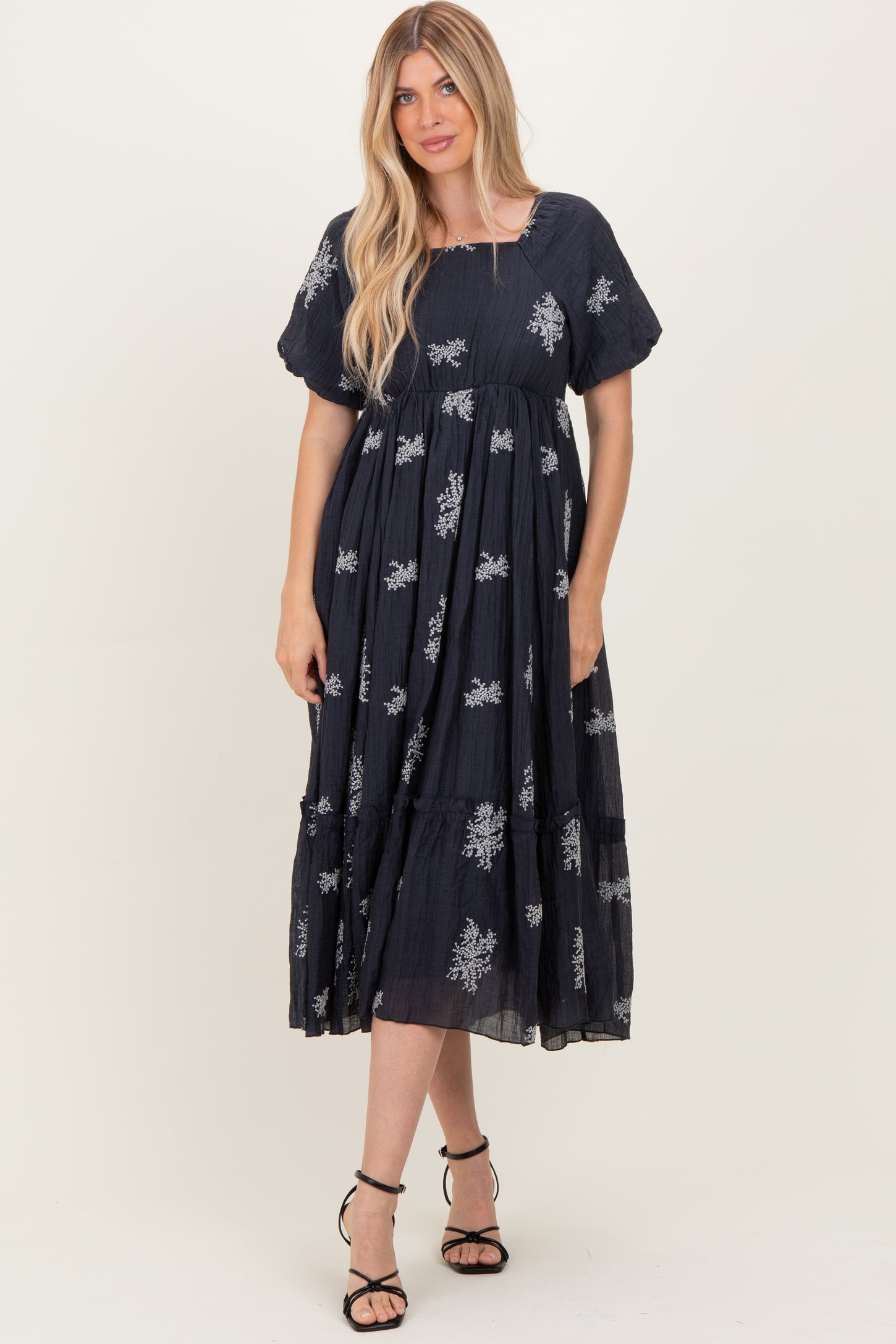 woman wearing a navy blue maxi dress with floral embroidery and puff sleeves, standing with hands in pockets