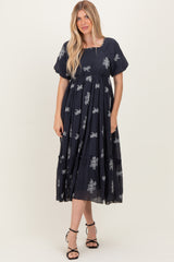 woman wearing a navy blue maxi dress with floral embroidery and puff sleeves, standing with hands in pockets