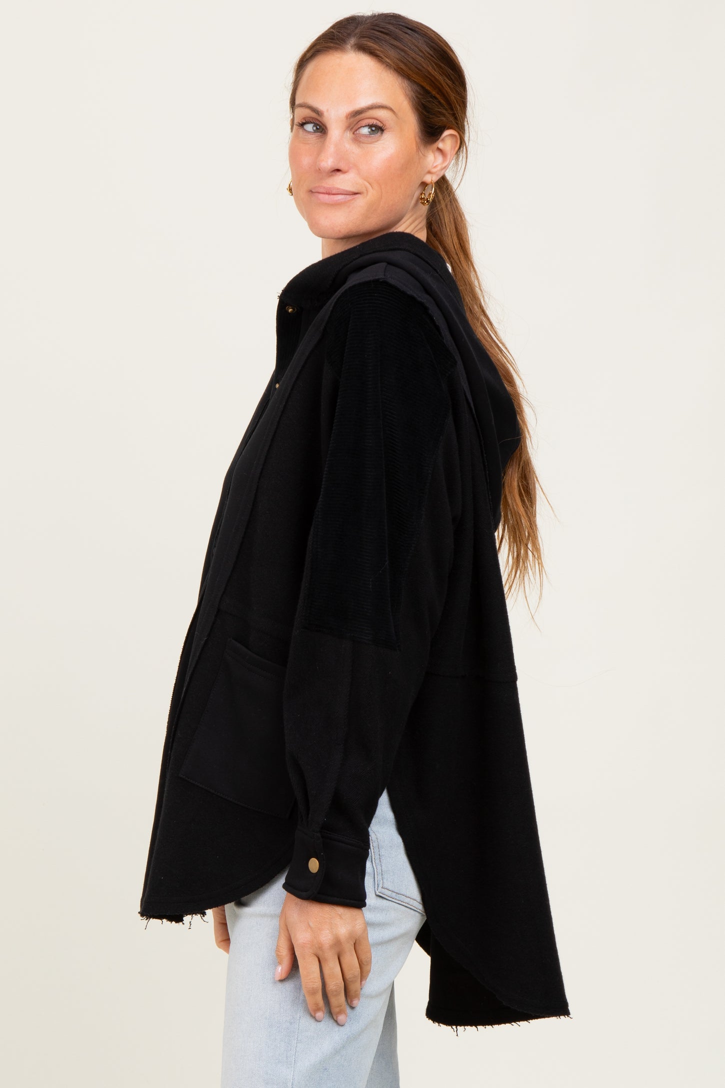 woman wearing a black hooded long sleeve shirt jacket with pockets and a curved hem, styled with light jeans