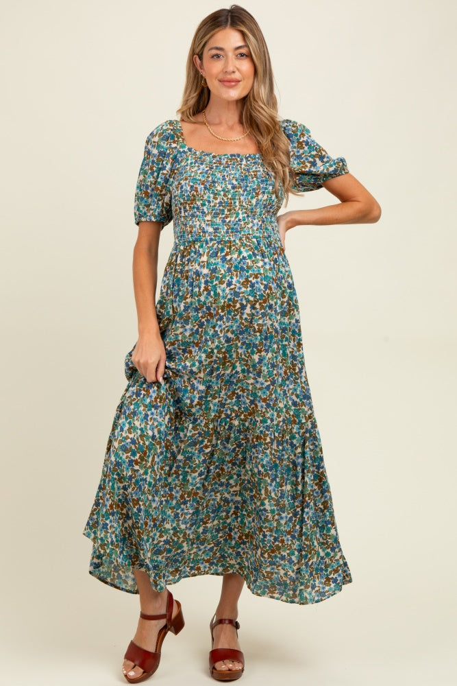 woman wearing a green floral print maternity dress with puff sleeves and a smocked bodice, holding the skirt with one hand