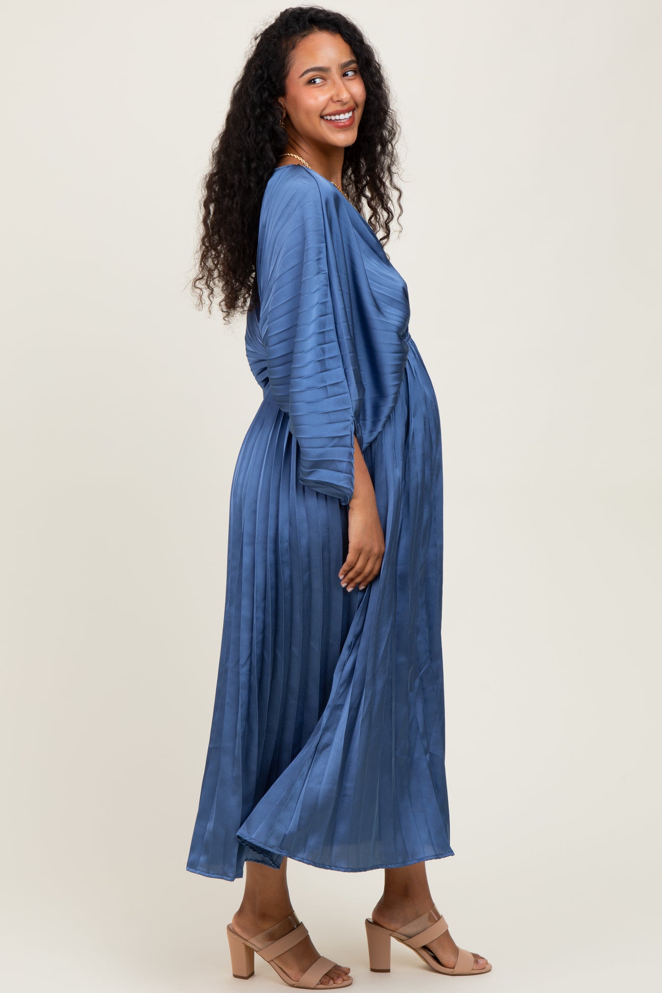 woman wearing a blue pleated maxi maternity dress, showcasing her baby bump and smiling at the camera