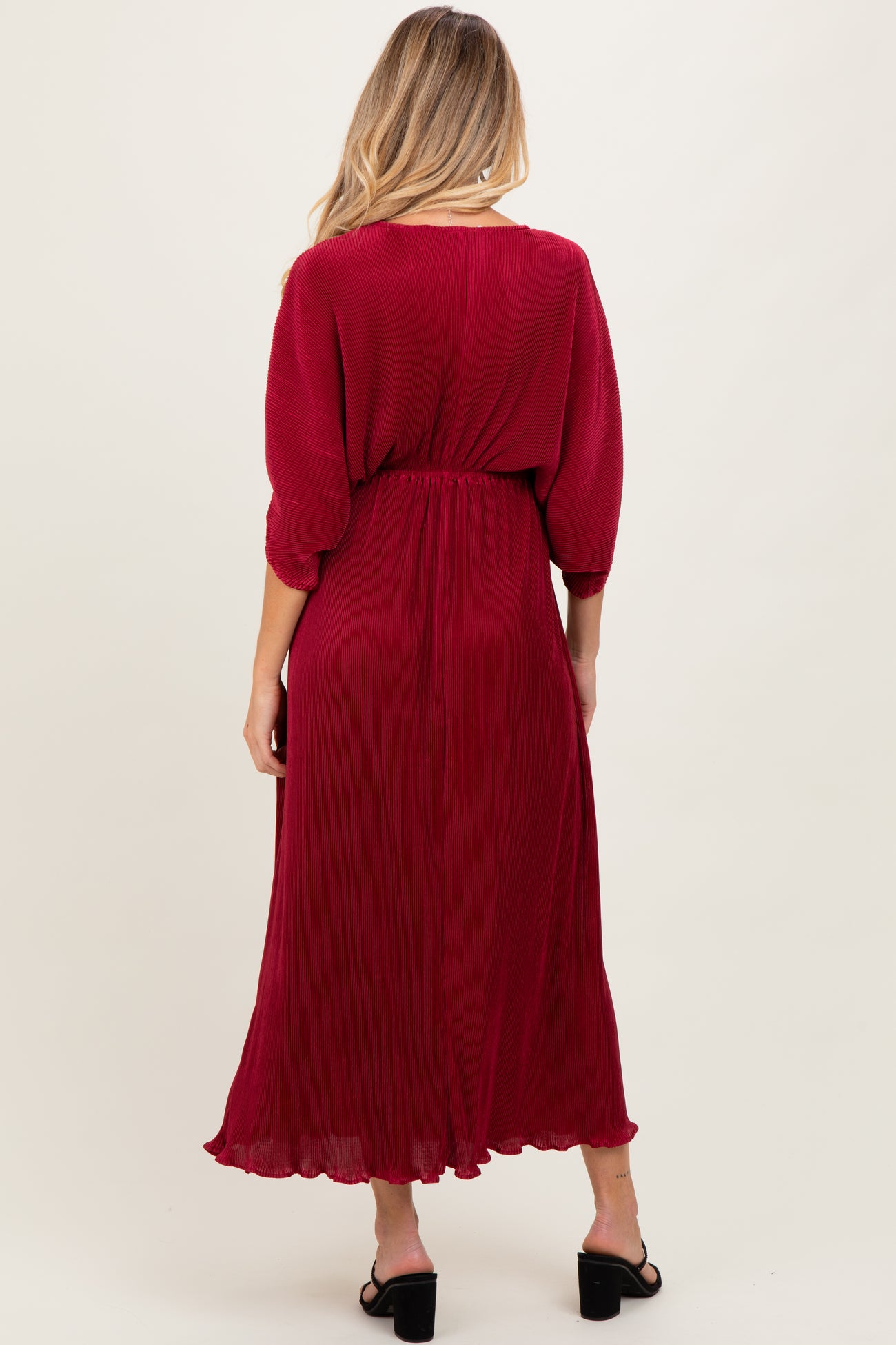 woman wearing a red pleated maternity maxi dress with a cinched waist and three-quarter sleeves, showing her baby bump from behind