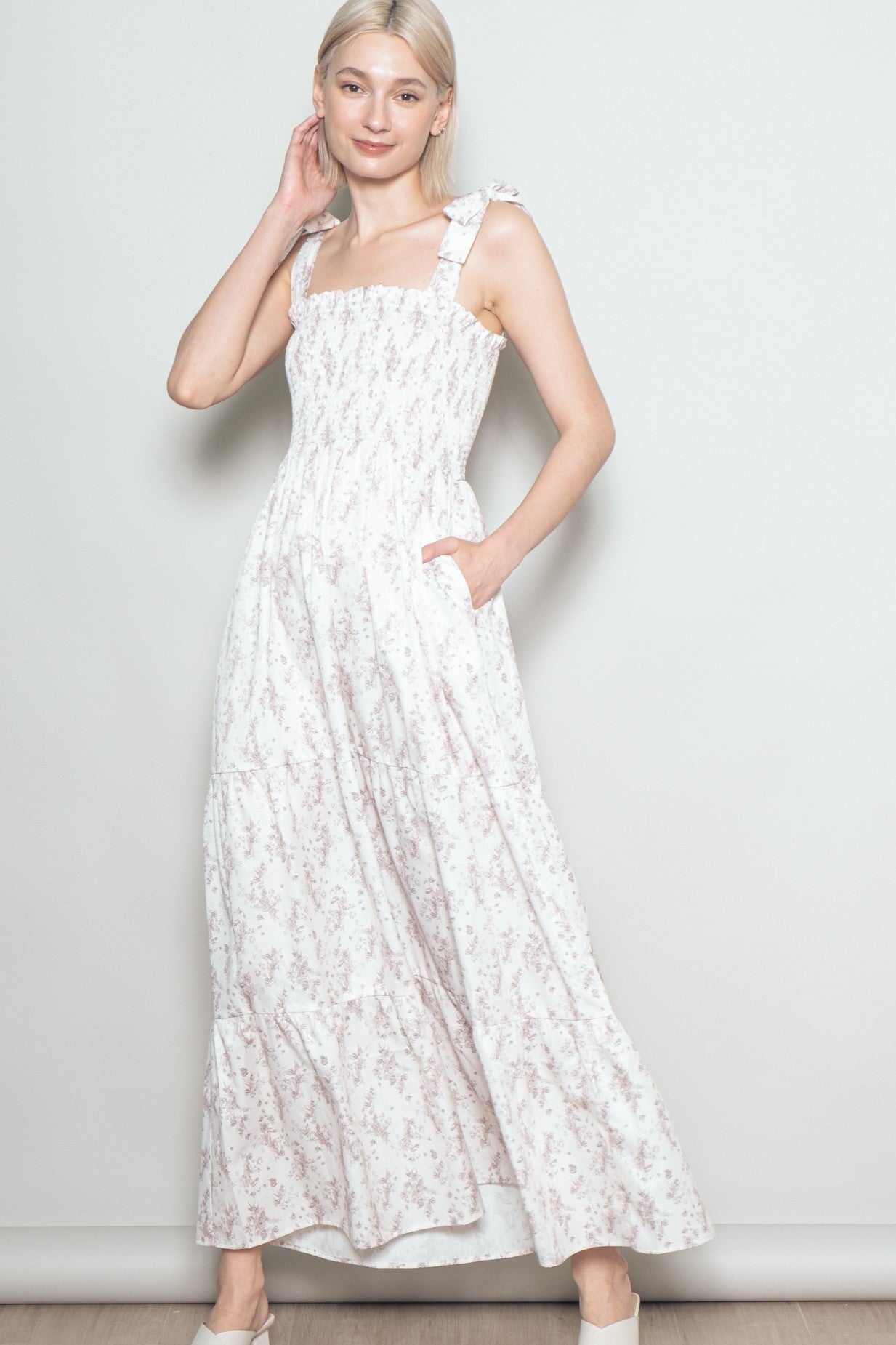 woman wearing a white maxi dress with a floral print, straps, and pockets, posing with one hand on her hip