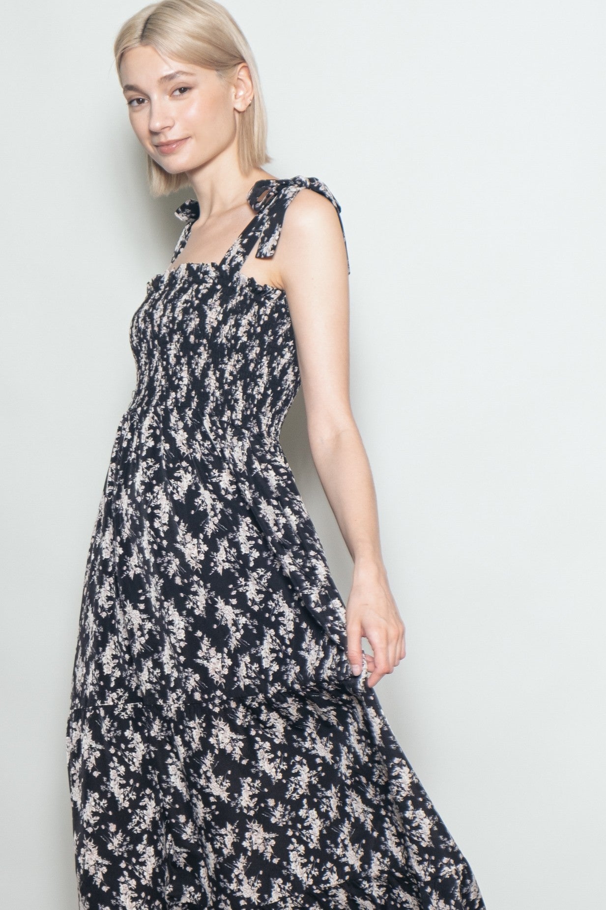 woman wearing a black floral maxi dress with smocked bodice and tie straps, standing in a neutral backdrop