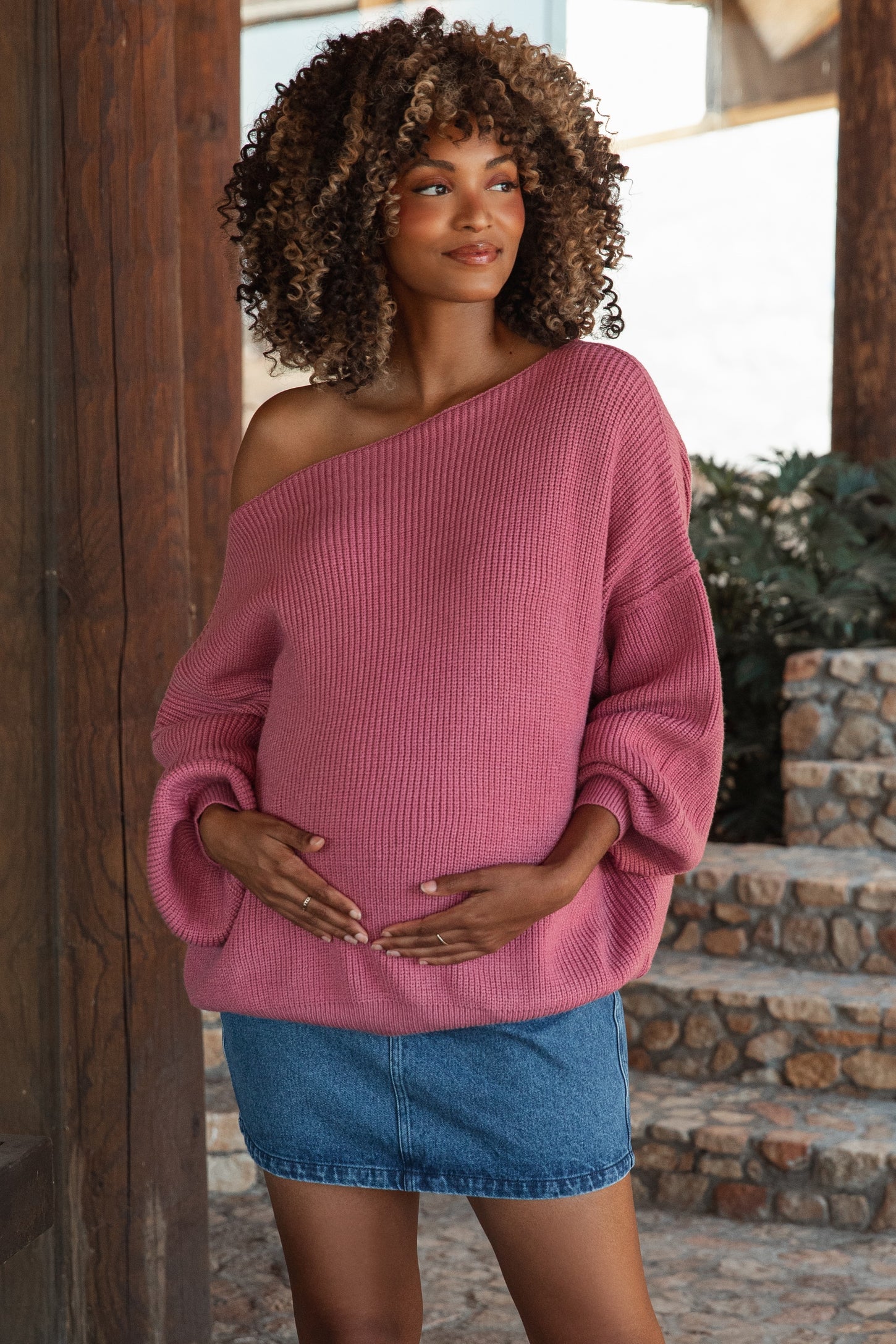 woman wearing a pink off-the-shoulder maternity sweater and a denim skirt, gently cradling her baby bump