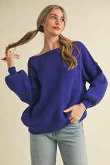 woman wearing a purple oversized ribbed sweater with off-the-shoulder neckline and balloon sleeves paired with light denim jeans