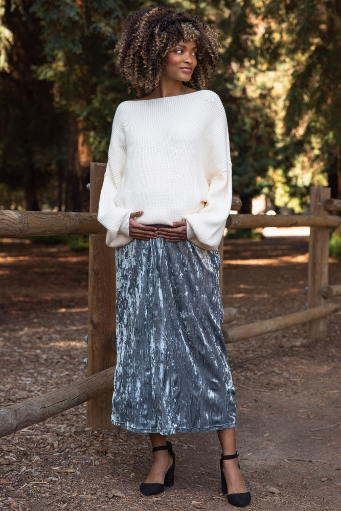 woman wearing a cream oversized sweater and a gray velvet maxi skirt, cradling her baby bump in a natural setting