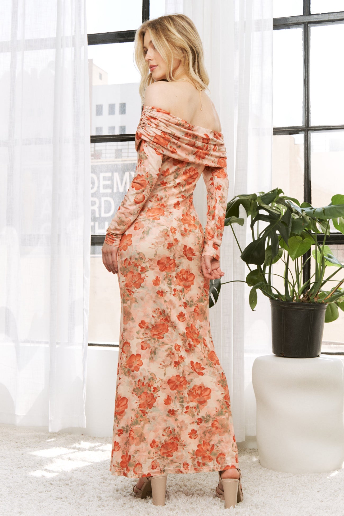 woman wearing a floral off-the-shoulder maxi dress with long sleeves, standing in front of a window