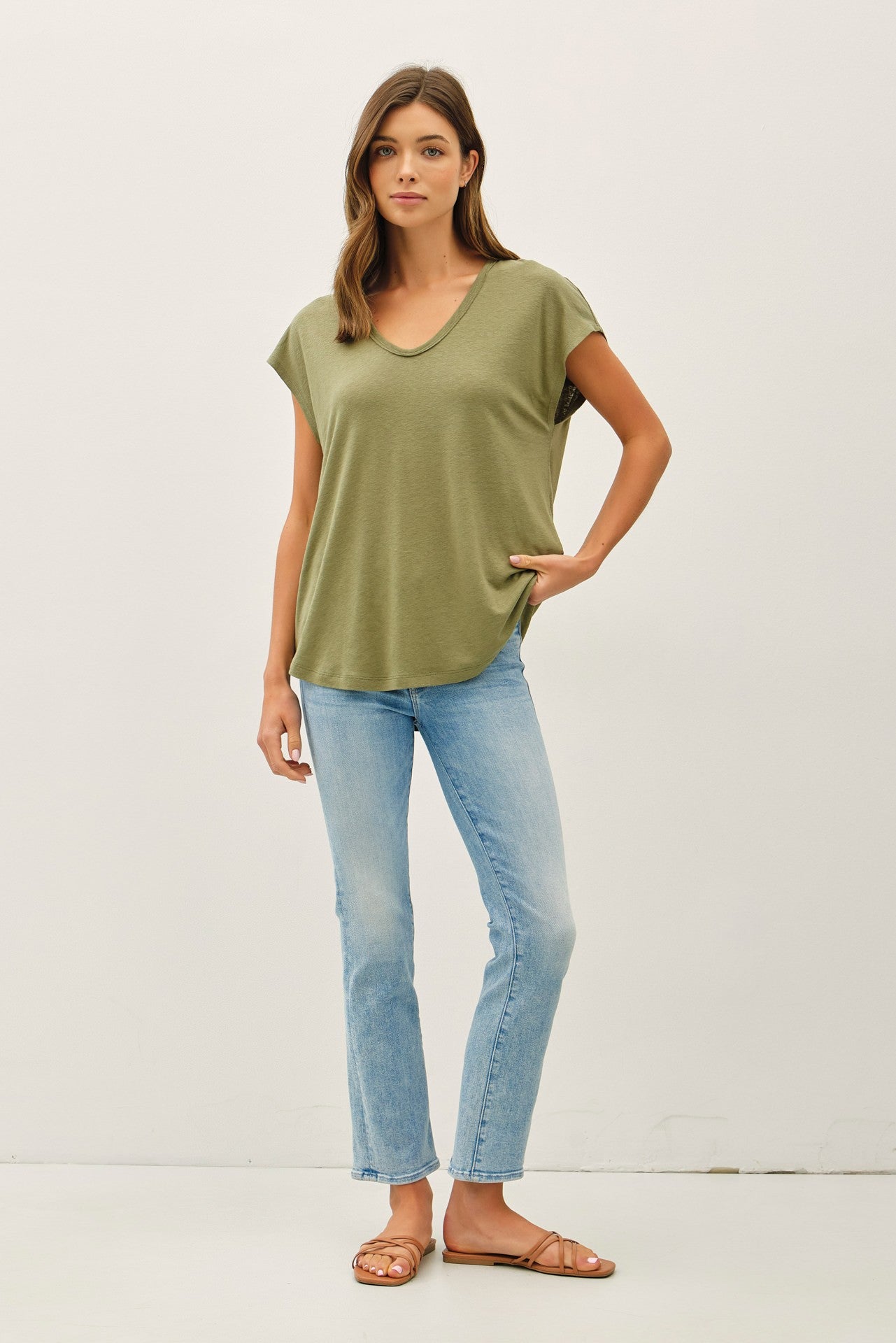 woman wearing an olive green short sleeve top and light blue flared jeans, standing with one hand on her hip