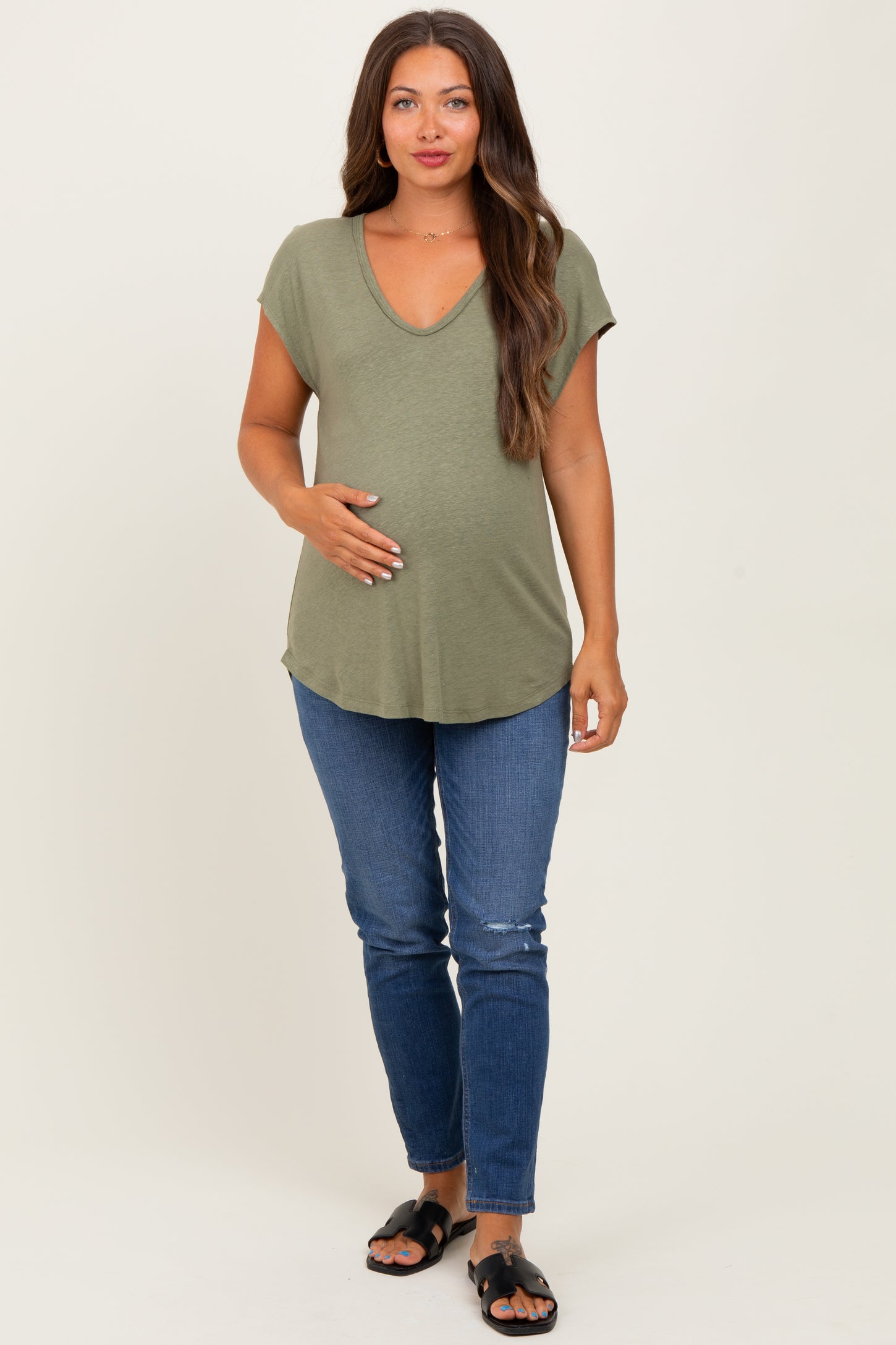 woman wearing an olive green maternity short sleeve top and holding her baby bump with blue jeans and sandals