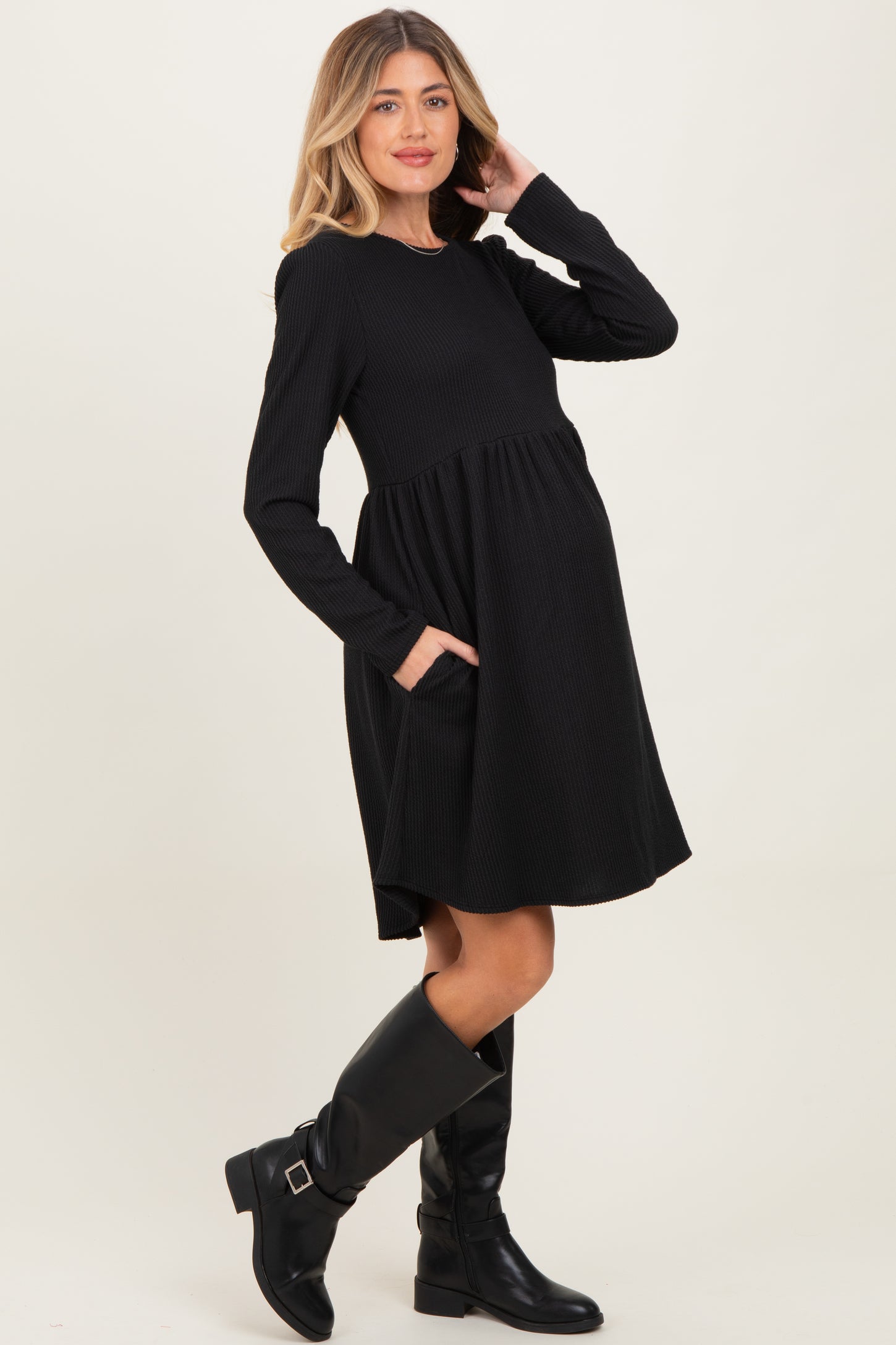 woman wearing a black long sleeve maternity dress with pockets and a baby bump, styled with knee-high black boots