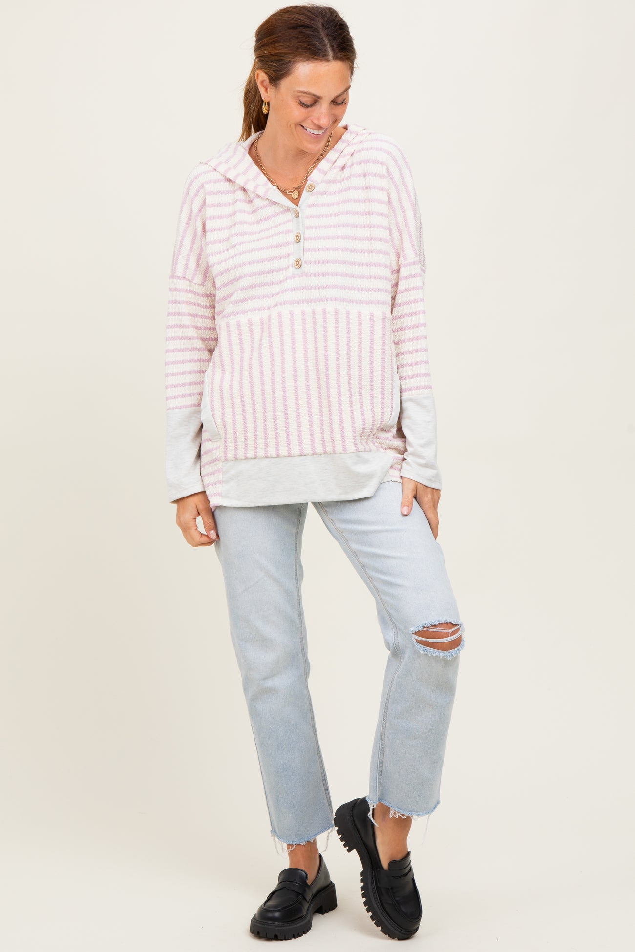 woman wearing a pink and white striped pullover hoodie with a button detail, paired with ripped light blue jeans and black slip-on shoes