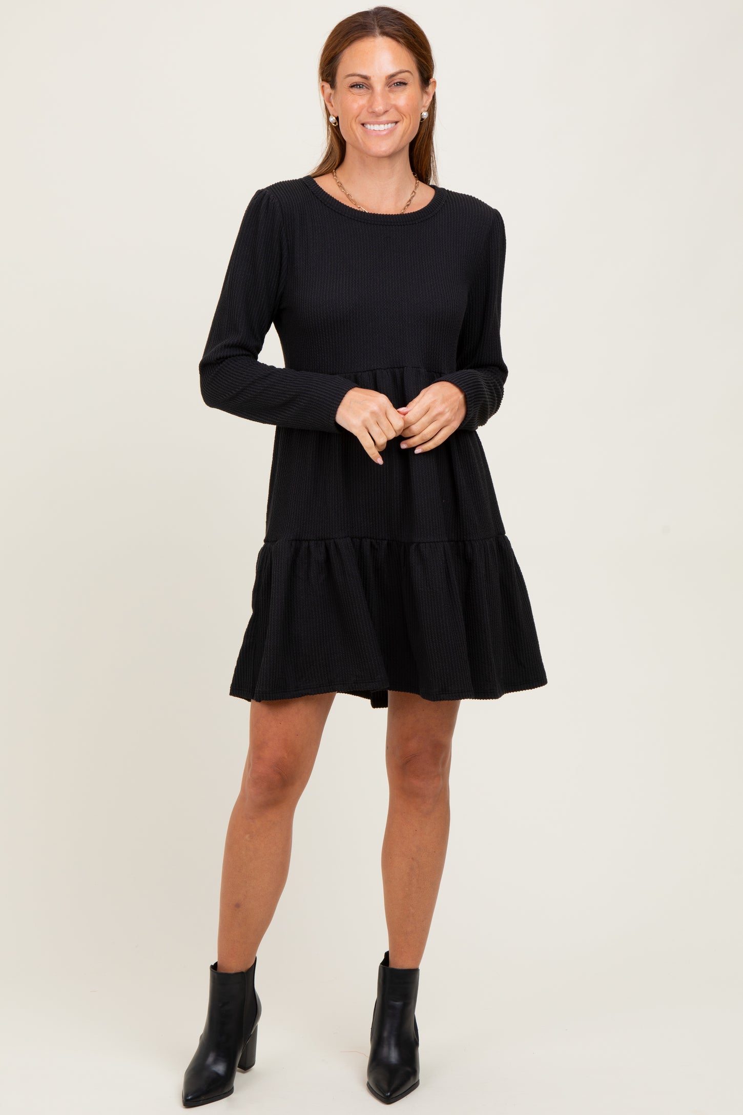 woman wearing a black long sleeve tiered mini dress and black ankle boots, smiling and standing against a plain background