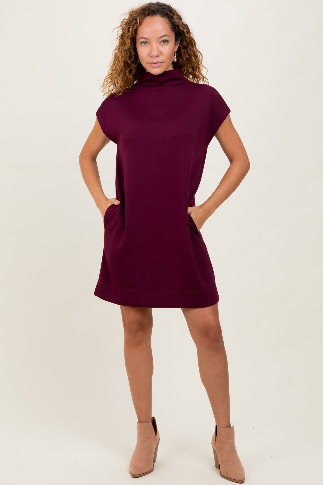 woman wearing a short burgundy dress with a cowl neck and side pockets, styled with beige ankle boots