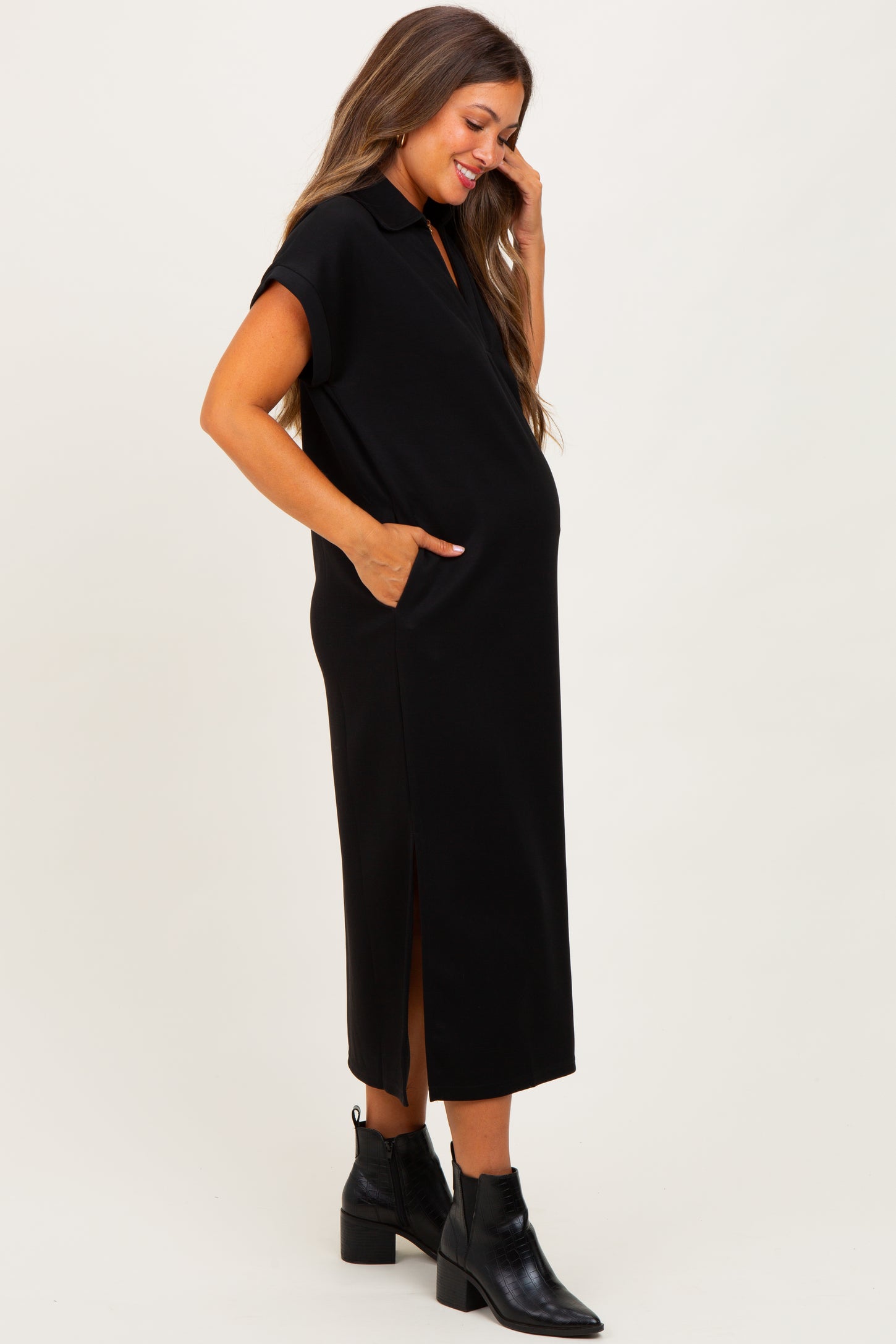 woman wearing a black maternity maxi dress with short sleeves and side pockets, smiling and showing her baby bump