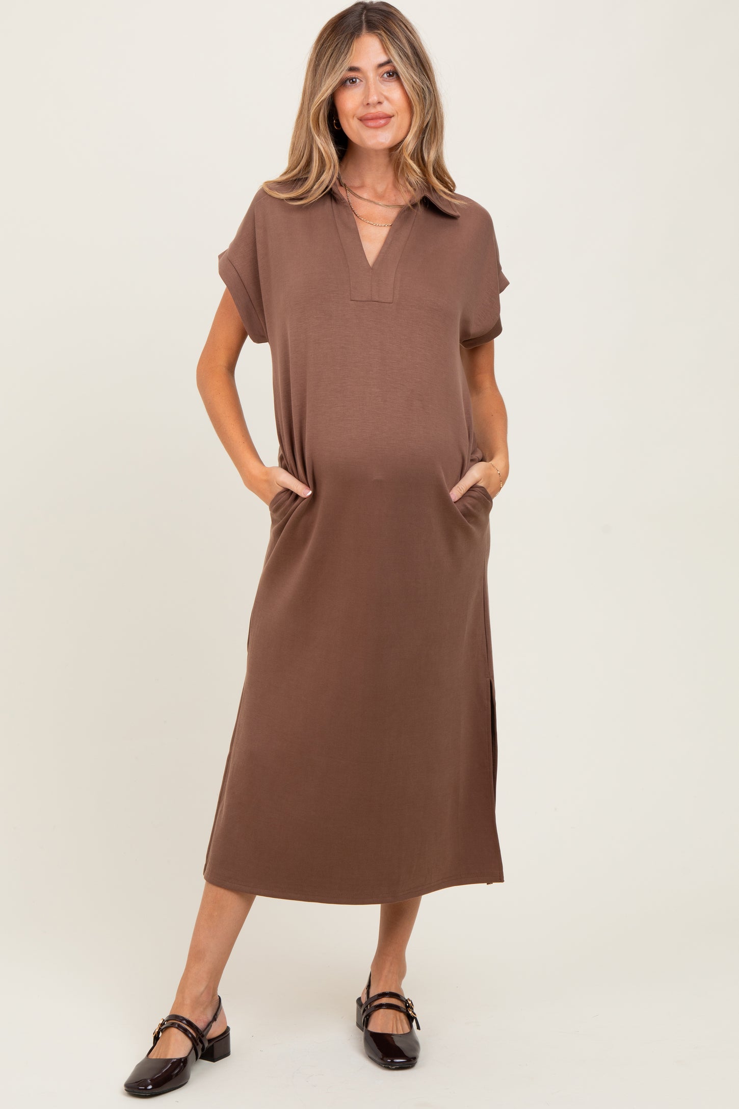woman wearing a brown short sleeve maternity dress with pockets and side slits, holding her baby bump