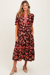 woman wearing a floral patterned brown maternity dress with a tiered skirt and red accents, posing with her hands in pockets