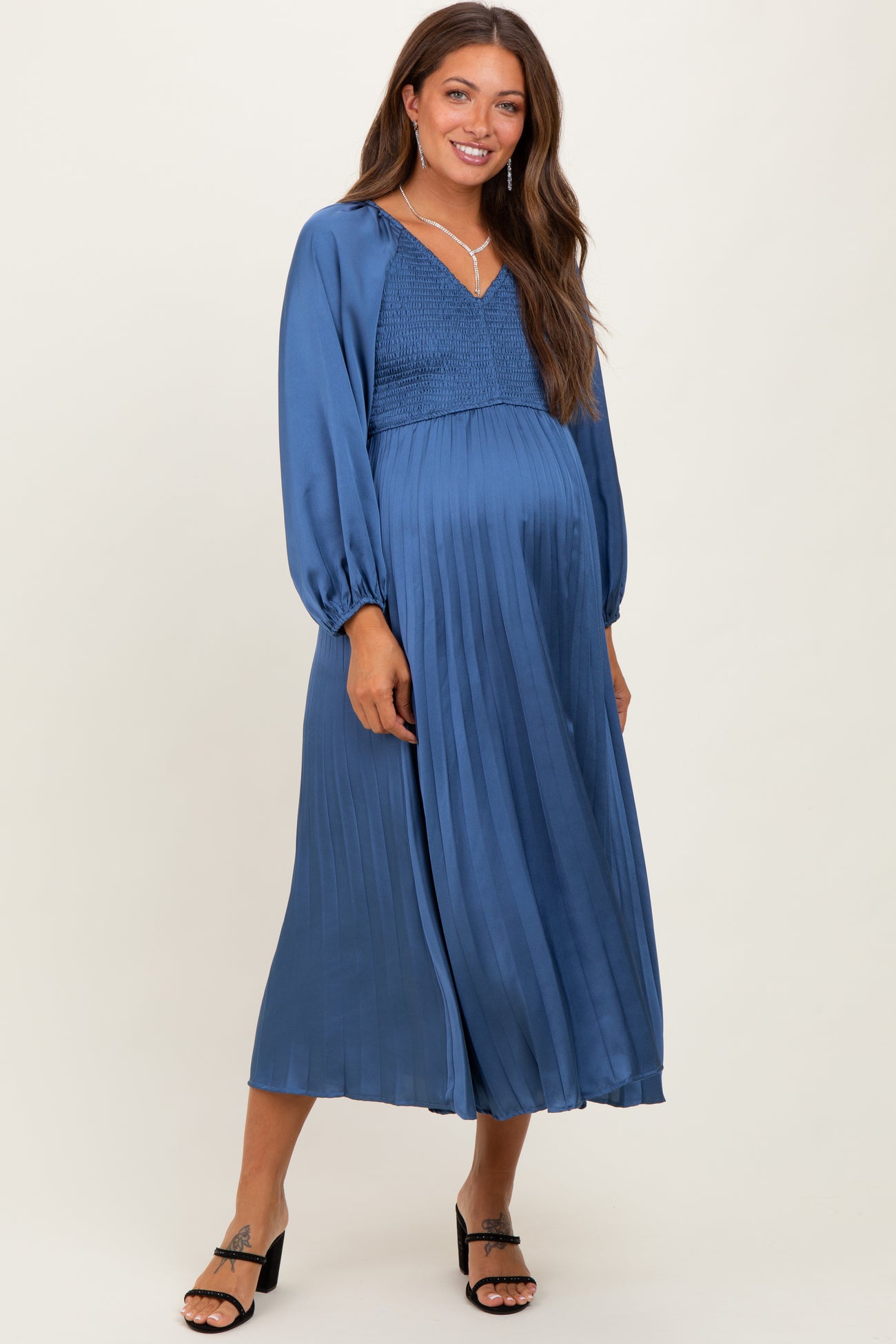 woman wearing a blue pleated maternity dress with a smocked bodice and long sleeves, showcasing her baby bump
