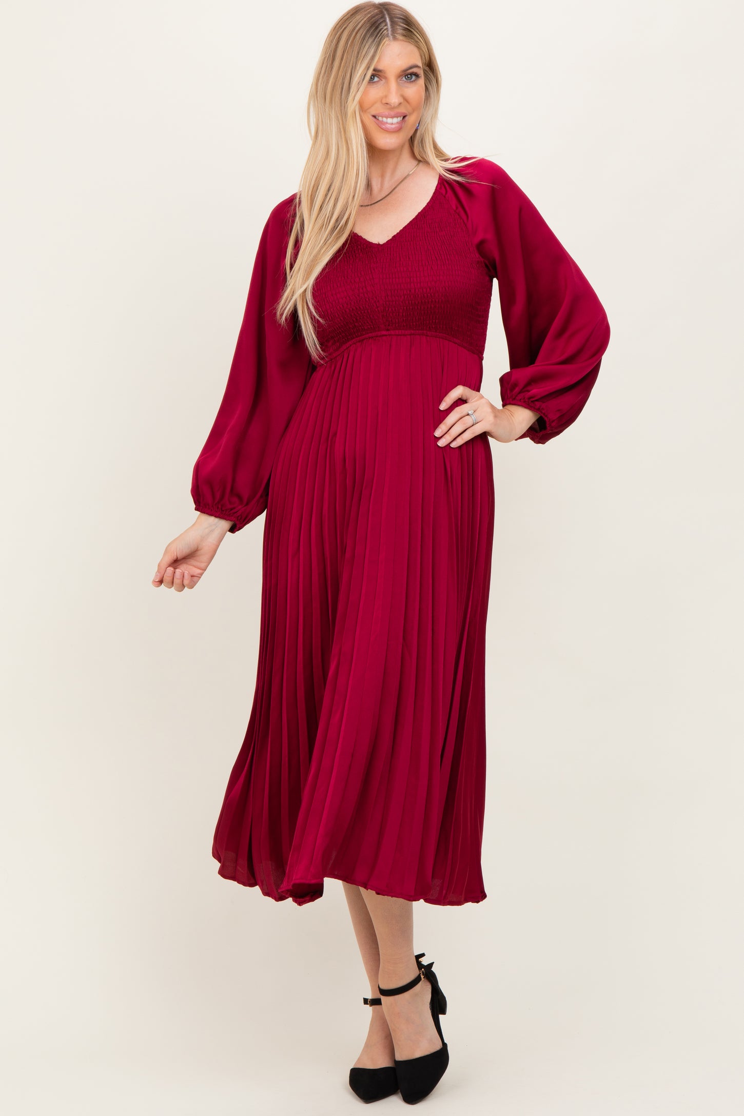 woman wearing a maroon pleated midi dress with a smocked bodice and long sleeves, styled with black heels