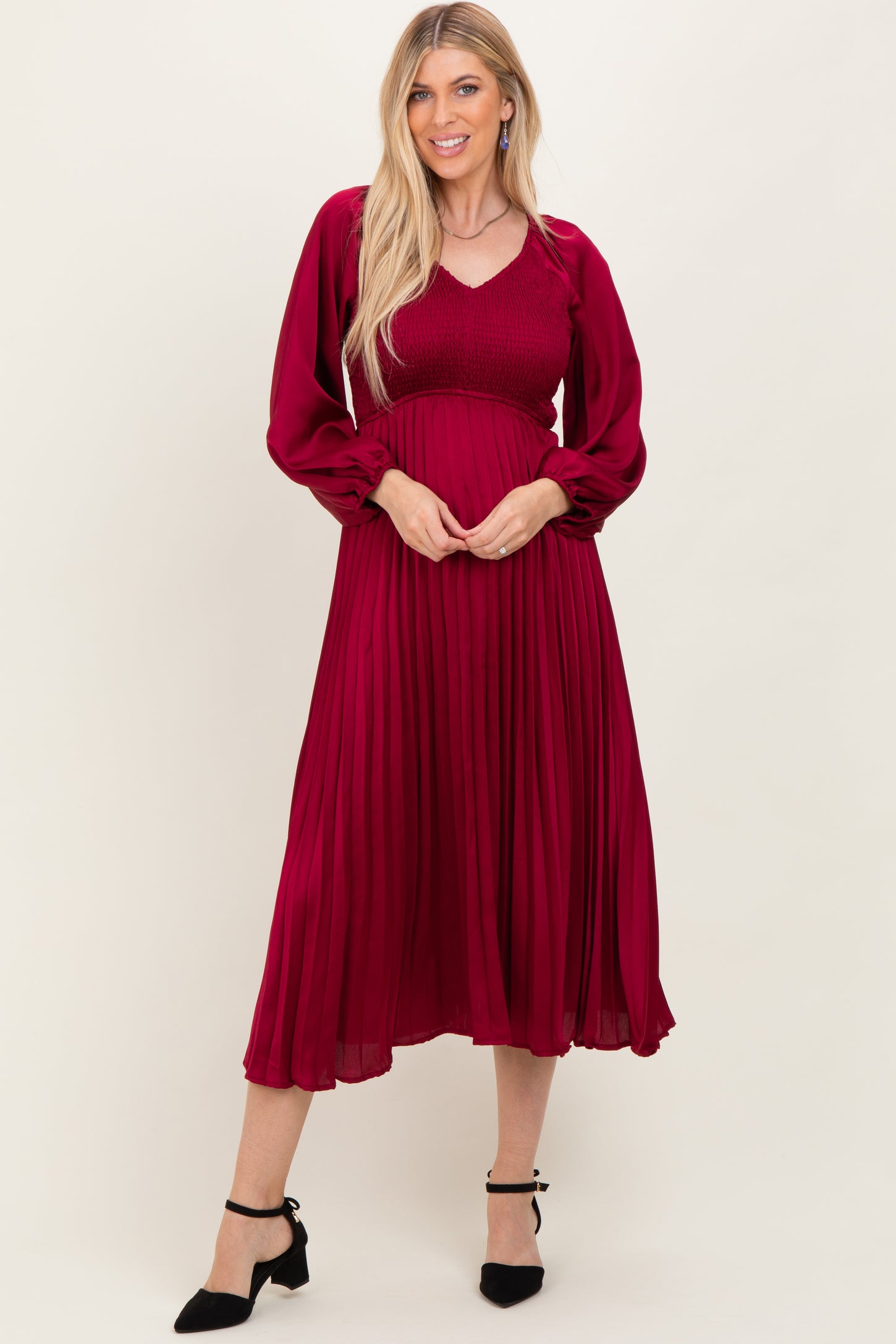 woman wearing a long sleeve burgundy pleated dress with a smocked bodice and flared skirt, posing with hands on her hips