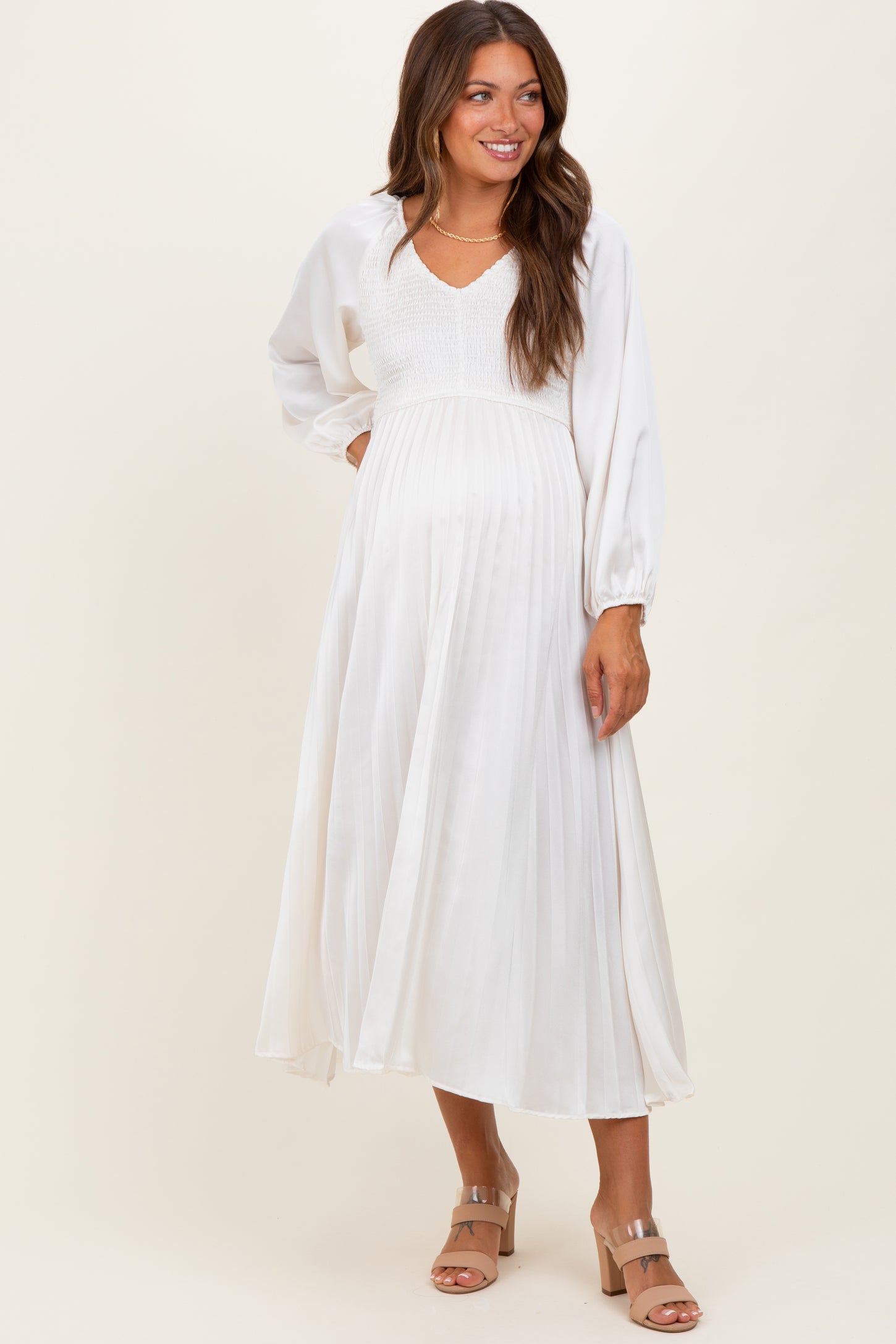 woman wearing a white pleated maternity dress with a v-neck and long sleeves, and showing her baby bump