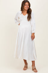 woman wearing a white pleated maternity dress with a v-neck and long sleeves, and showing her baby bump