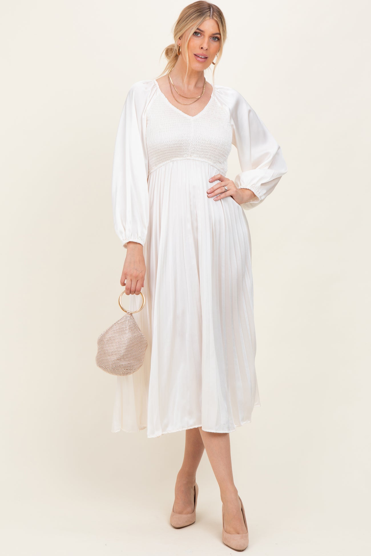 woman wearing a cream long sleeve pleated dress with a smocked bodice, holding a woven handbag and wearing heels