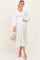woman wearing a cream long sleeve pleated dress with a smocked bodice, holding a woven handbag and wearing heels