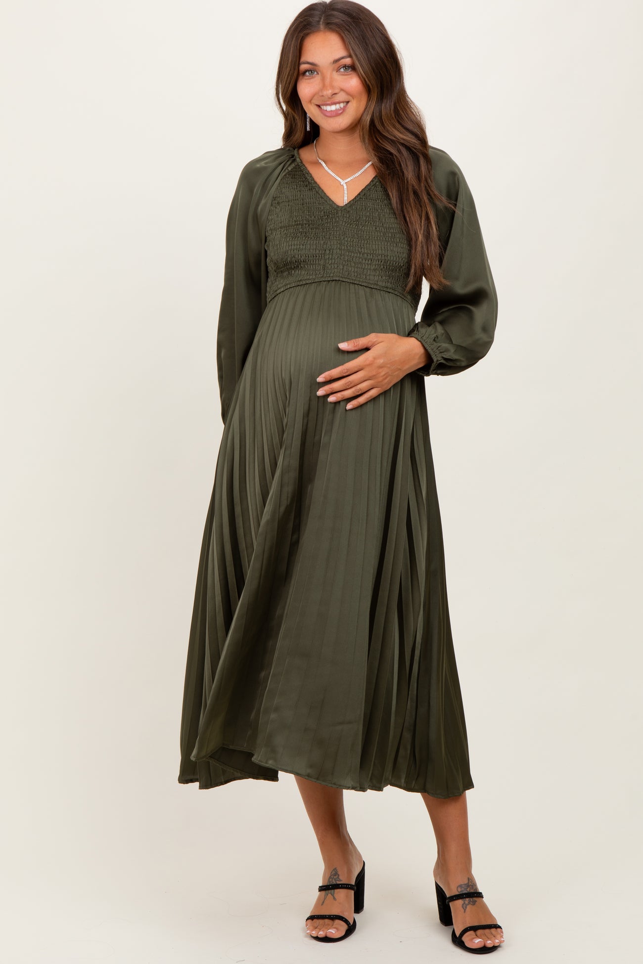 woman wearing an olive green pleated maternity dress with a smocked bodice and long sleeves, holding her baby bump