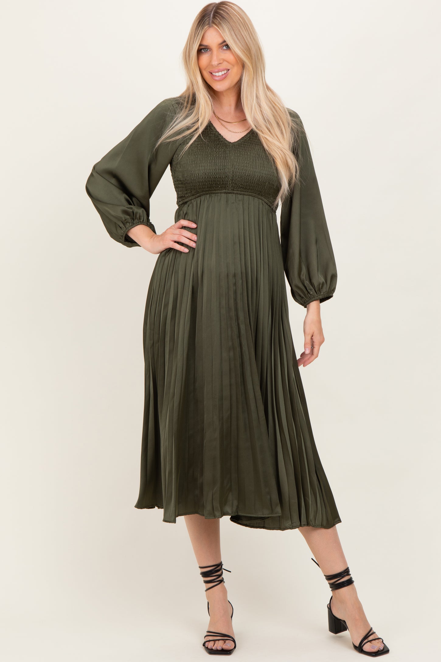 woman wearing an olive green pleated midi dress with long sleeves and a smocked bodice, styled with strappy heels