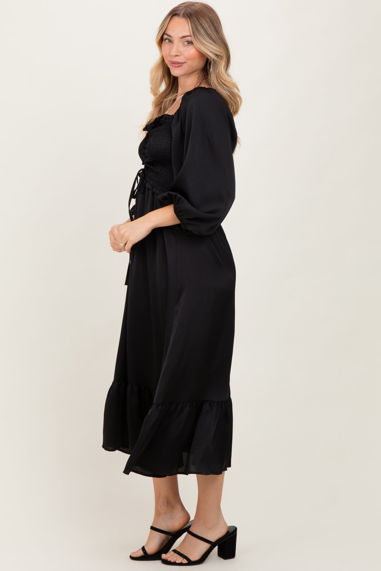 woman wearing a black smocked maxi dress with puff sleeves and a ruffled hem, posed sideways against a neutral background
