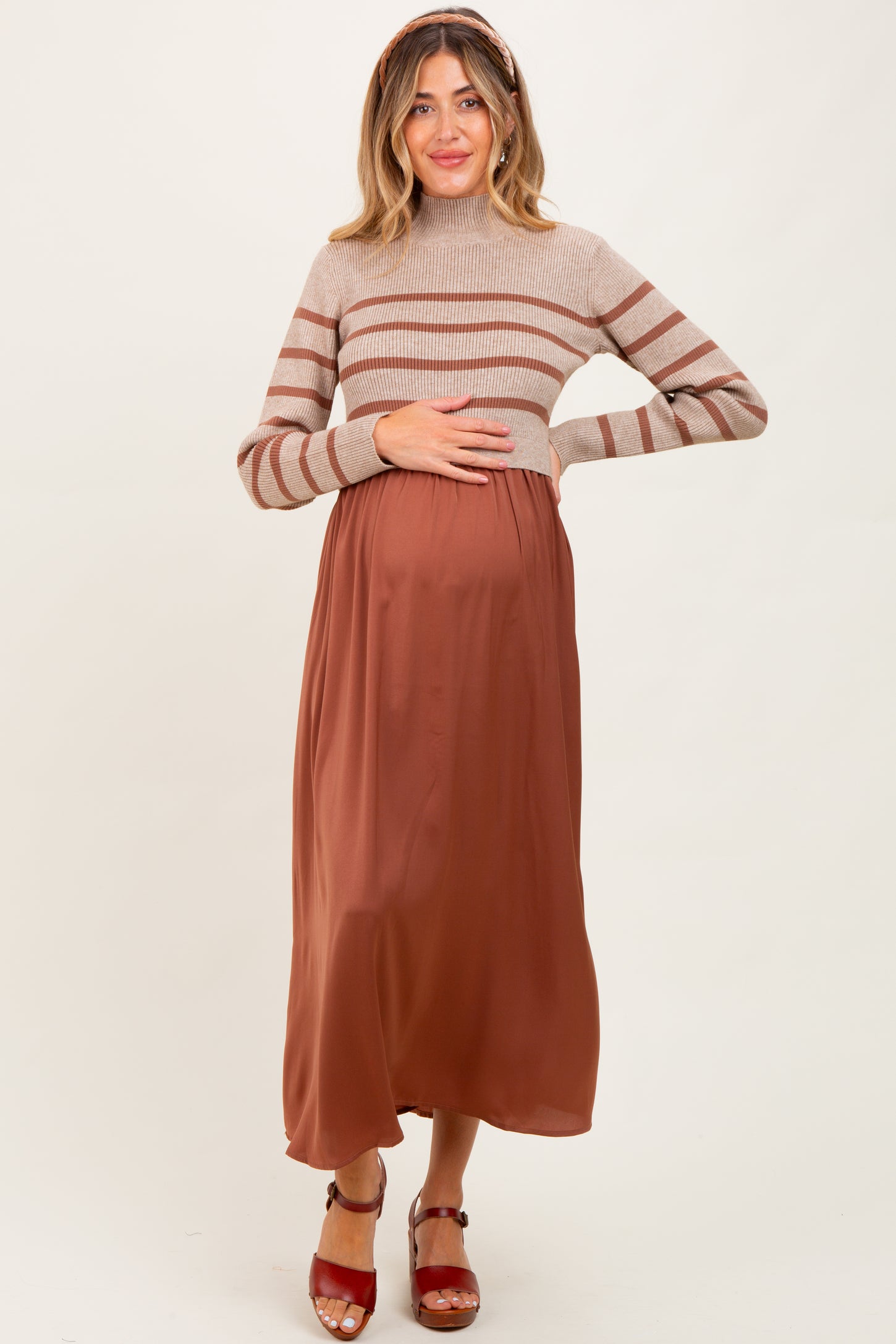 woman wearing a beige striped maternity sweater and a brown satin maternity dress, cradling her baby bump