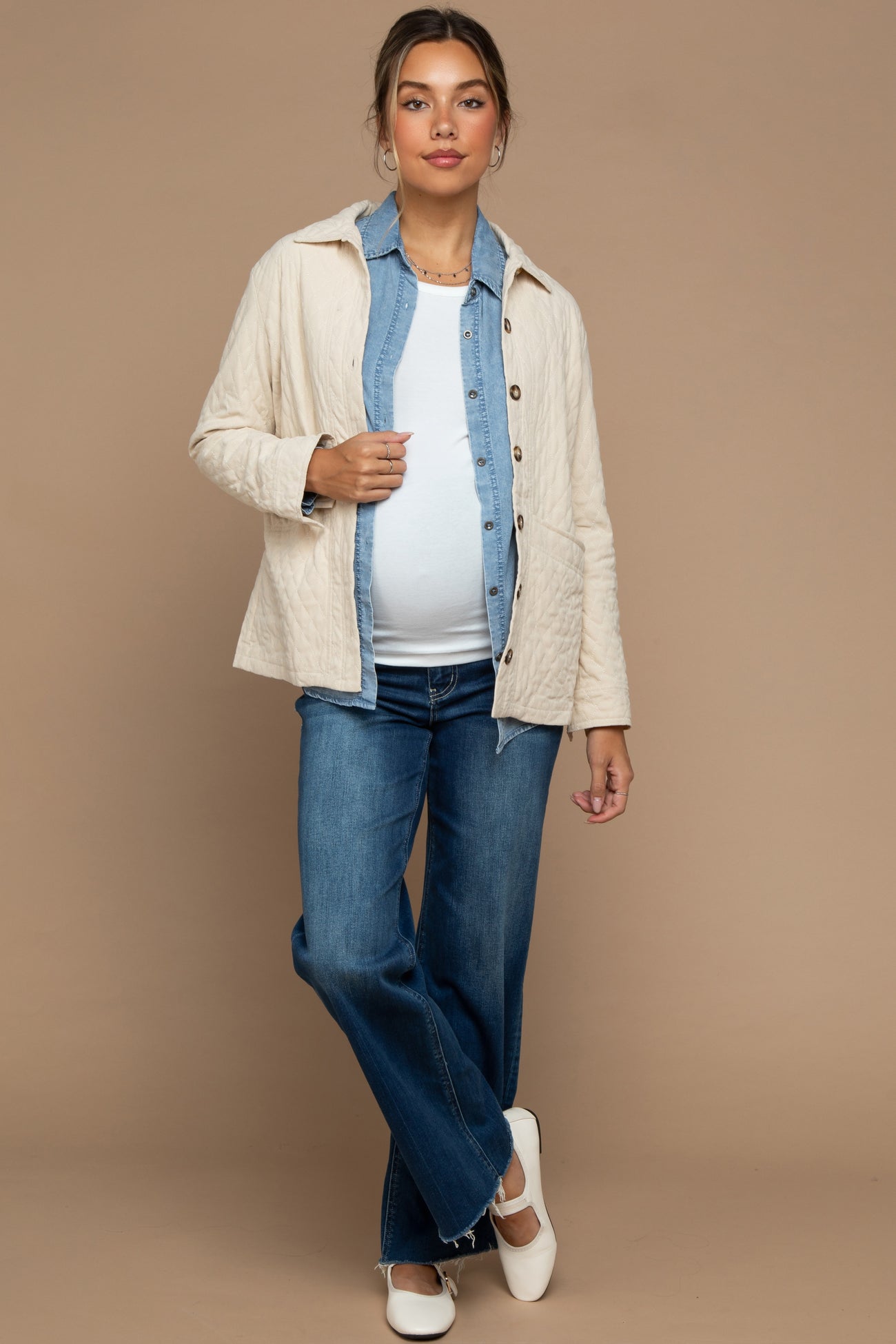 woman wearing a cream quilted maternity jacket over a denim shirt, white top, and wide-leg jeans, showing her baby bump