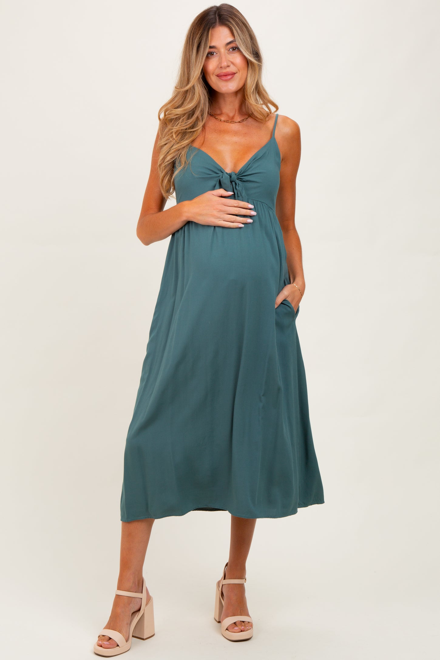 woman wearing a dark teal maternity dress with adjustable straps and a tie front, holding her baby bump