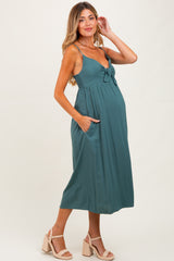 woman wearing a teal maternity dress with a bow detail and pockets, showing her baby bump