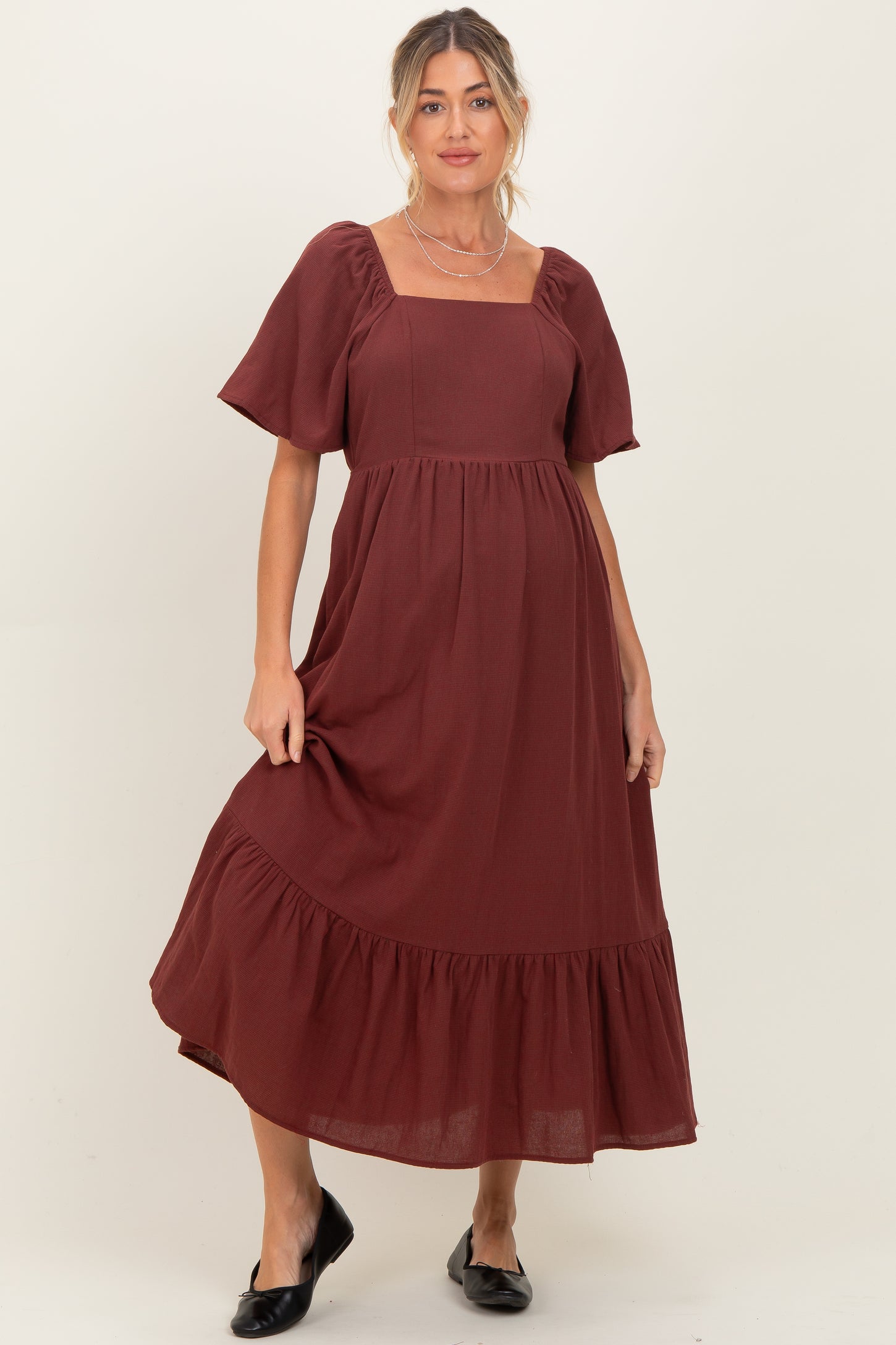 woman wearing a burgundy maternity dress with a square neckline and flutter sleeves, featuring a tiered hem