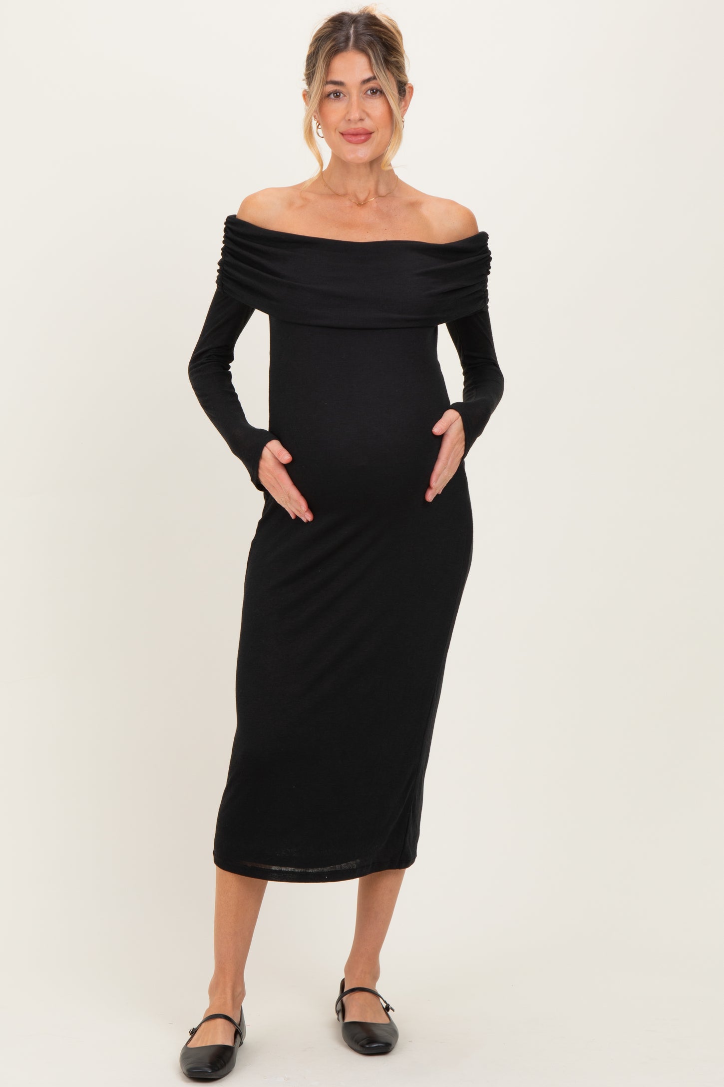 woman wearing a black off-the-shoulder maternity dress, holding her baby bump with a fitted silhouette