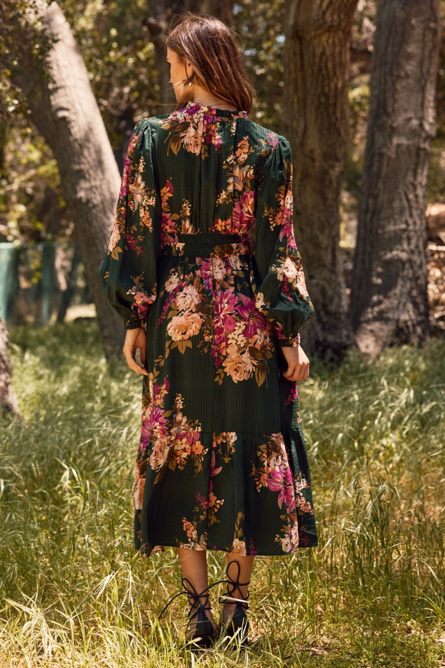 woman wearing a dark green floral maxi dress with long sleeves standing in a grassy area surrounded by trees